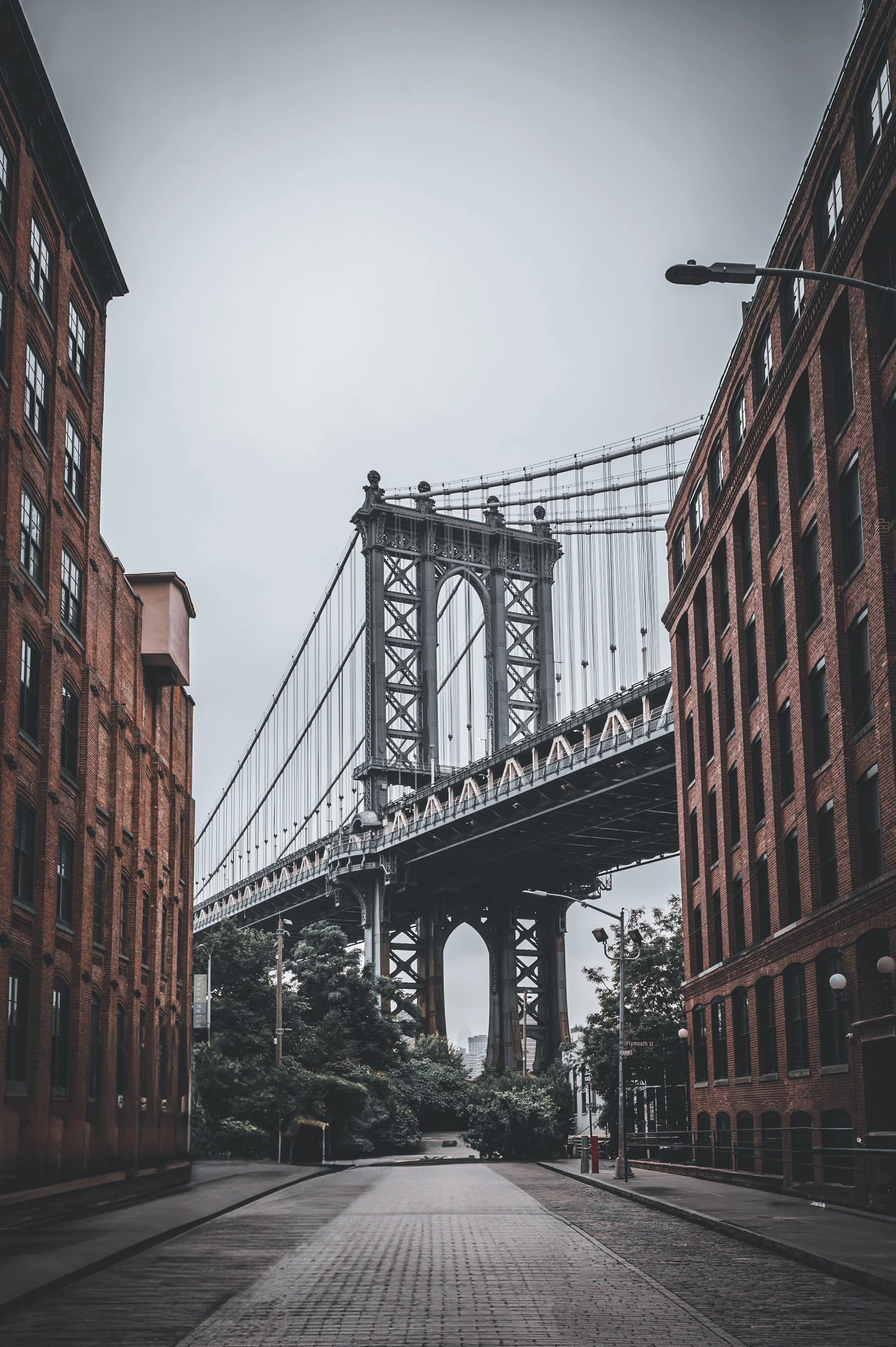 View of the Manhattan Bridge in Brooklyn, New York, through an empty cobblestone street flanked by red brick buildings on a cloudy day.