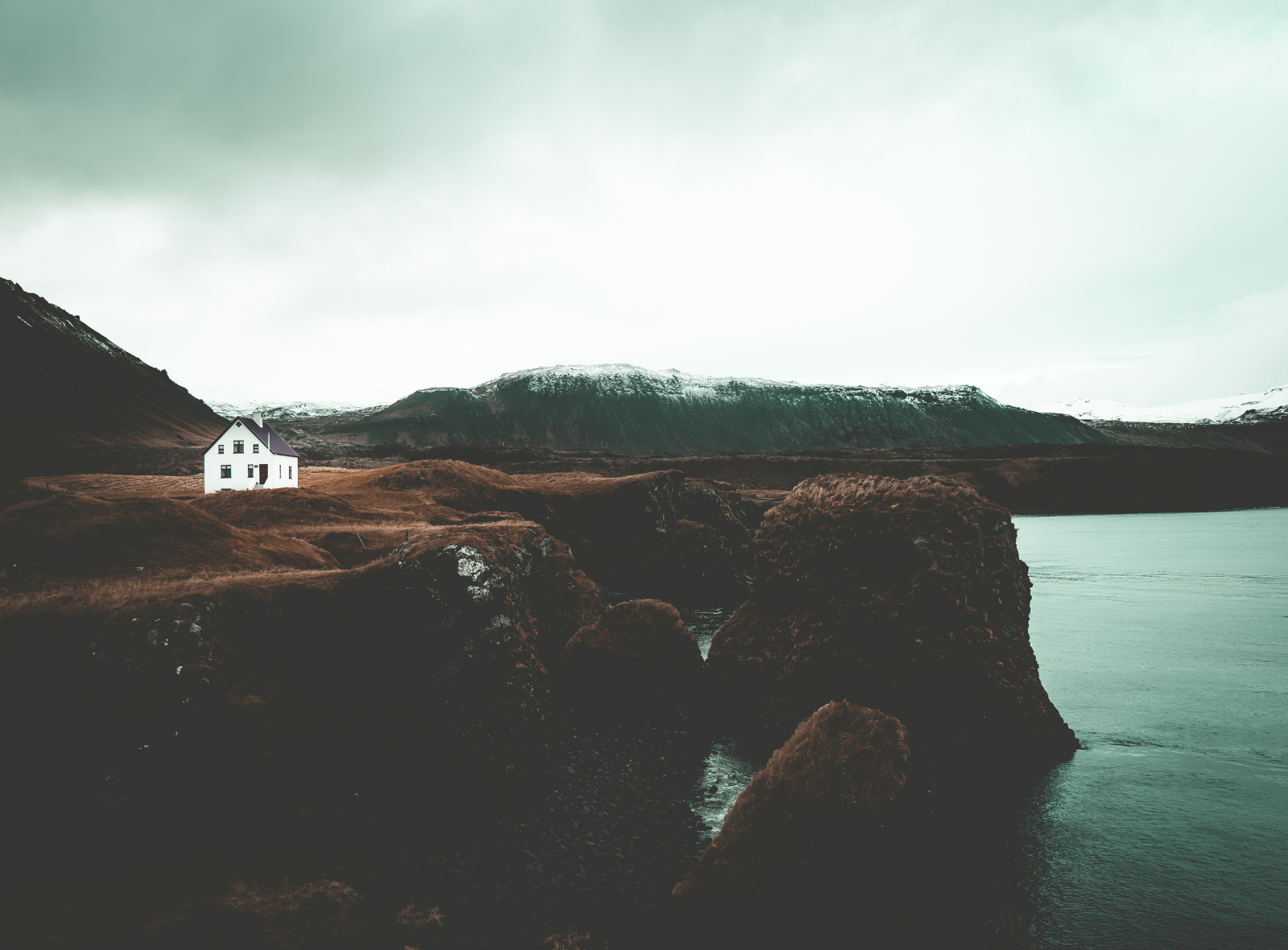 A white house with a dark roof situated on a grassy hill near a body of water, with mountains in the background under a cloudy sky. Iceland