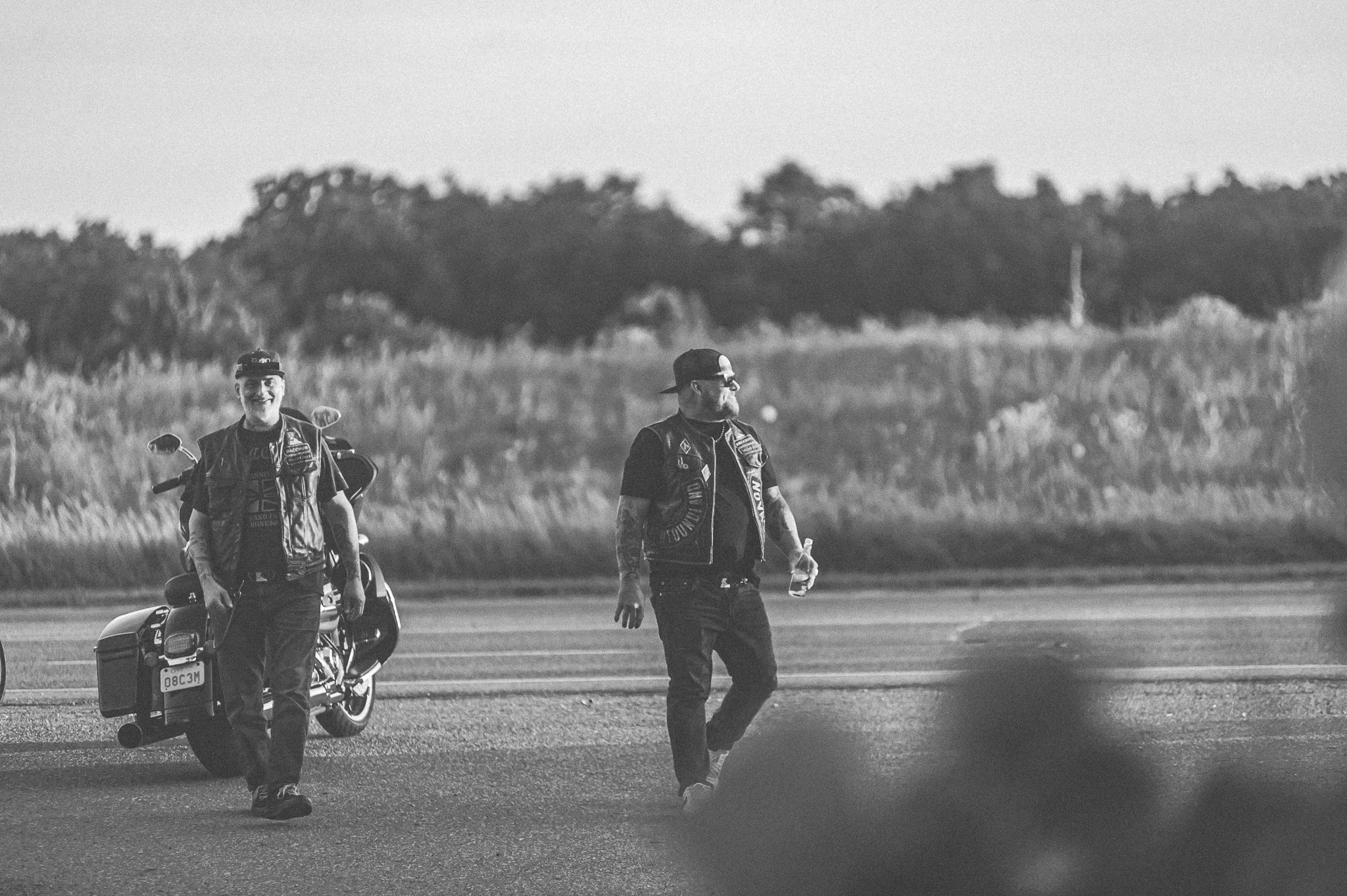 Two men dressed in motorcycle gear, one smiling and walking towards the camera with a motorcycle behind him, the other walking with sunglasses and tattoos, on an open road with grassy fields and trees in the background.