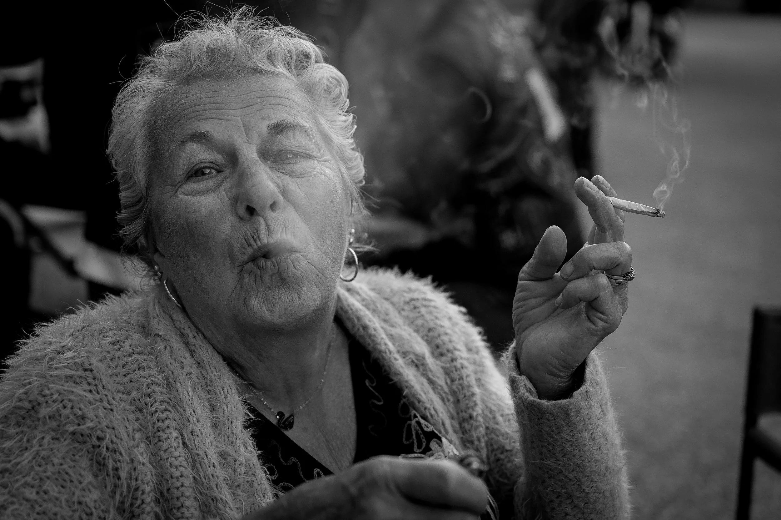 An elderly woman with curly hair blowing a kiss, holding a cigarette between her fingers, with smoke rising from it.