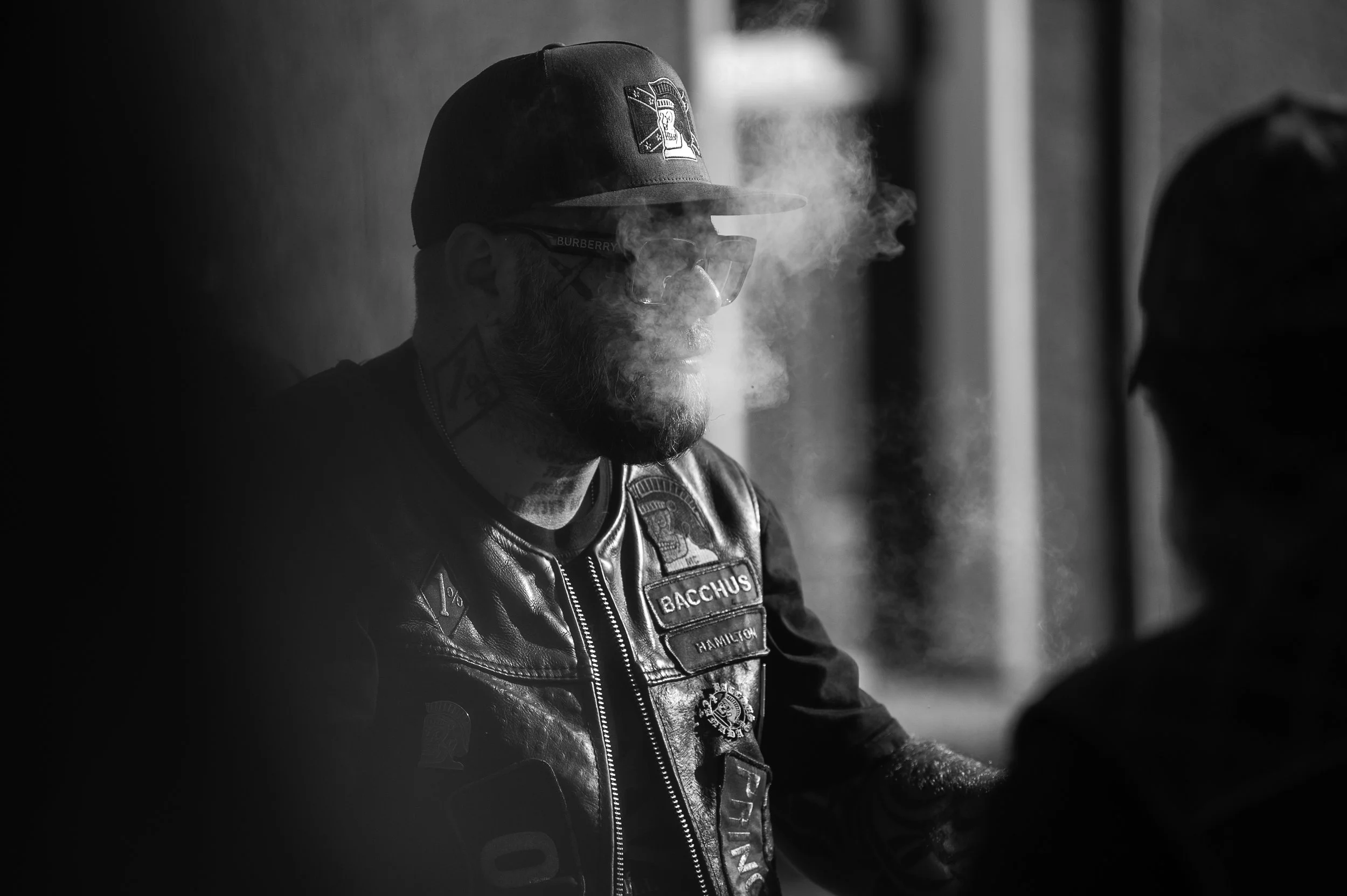 A man with a beard, wearing sunglasses, a cap, and a leather vest with patches, is exhaling smoke in a dimly lit setting.