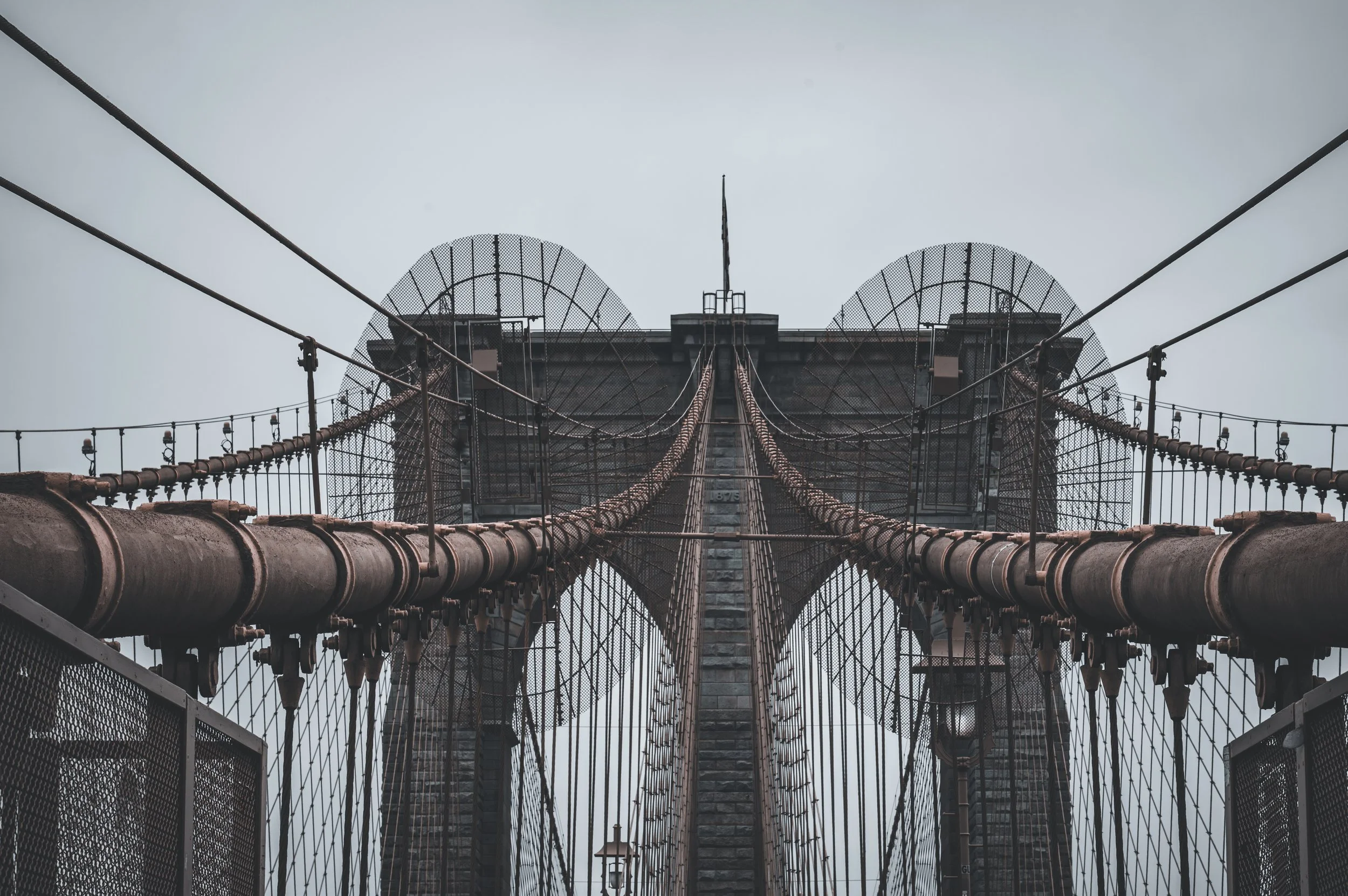 Looking up at the Brooklyn Bridge from the pedestrian walkway, showing the bridge's cables and stone towers against a cloudy sky.