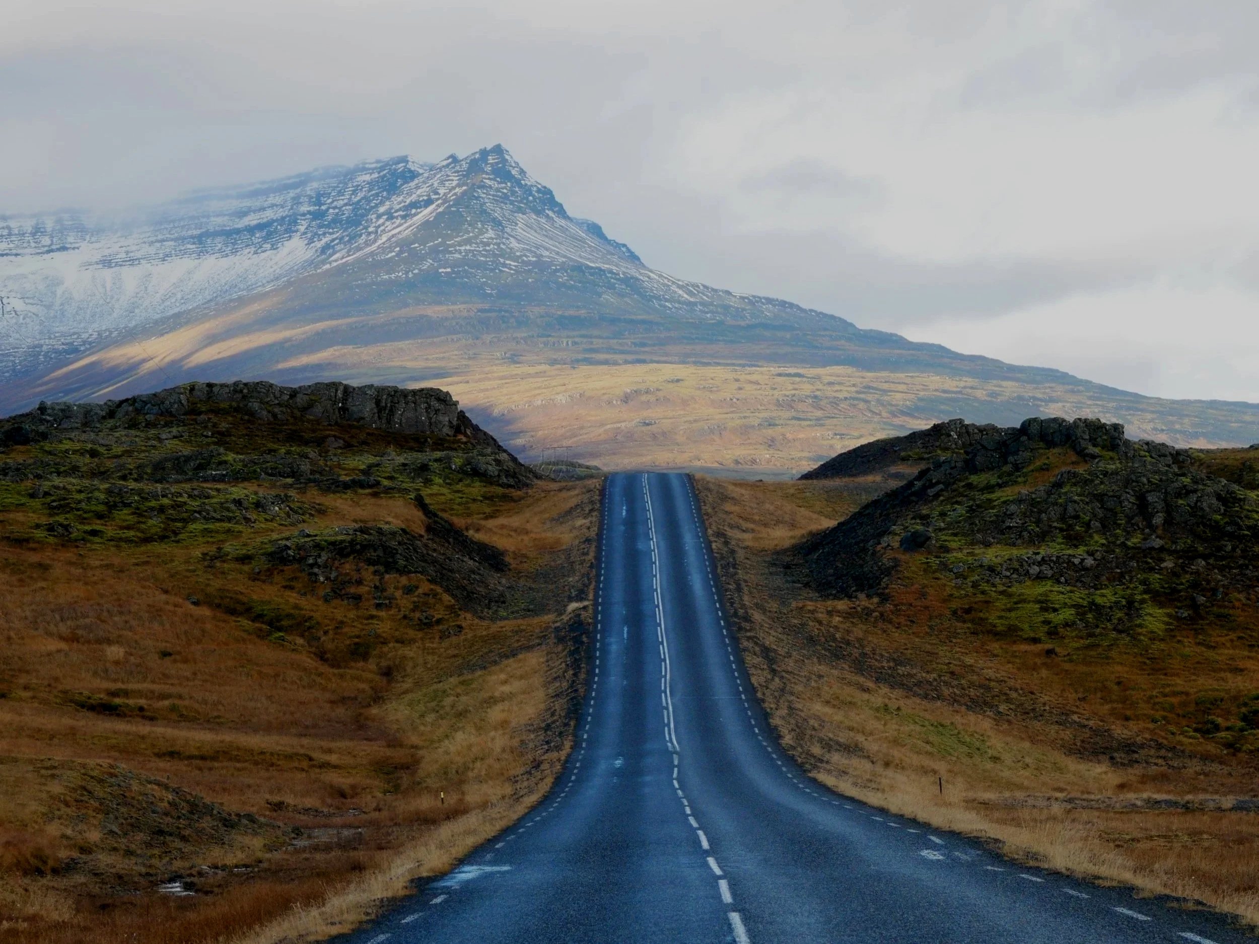 A winding road leading towards a mountain with snow-capped peaks against a cloudy sky. Iceland