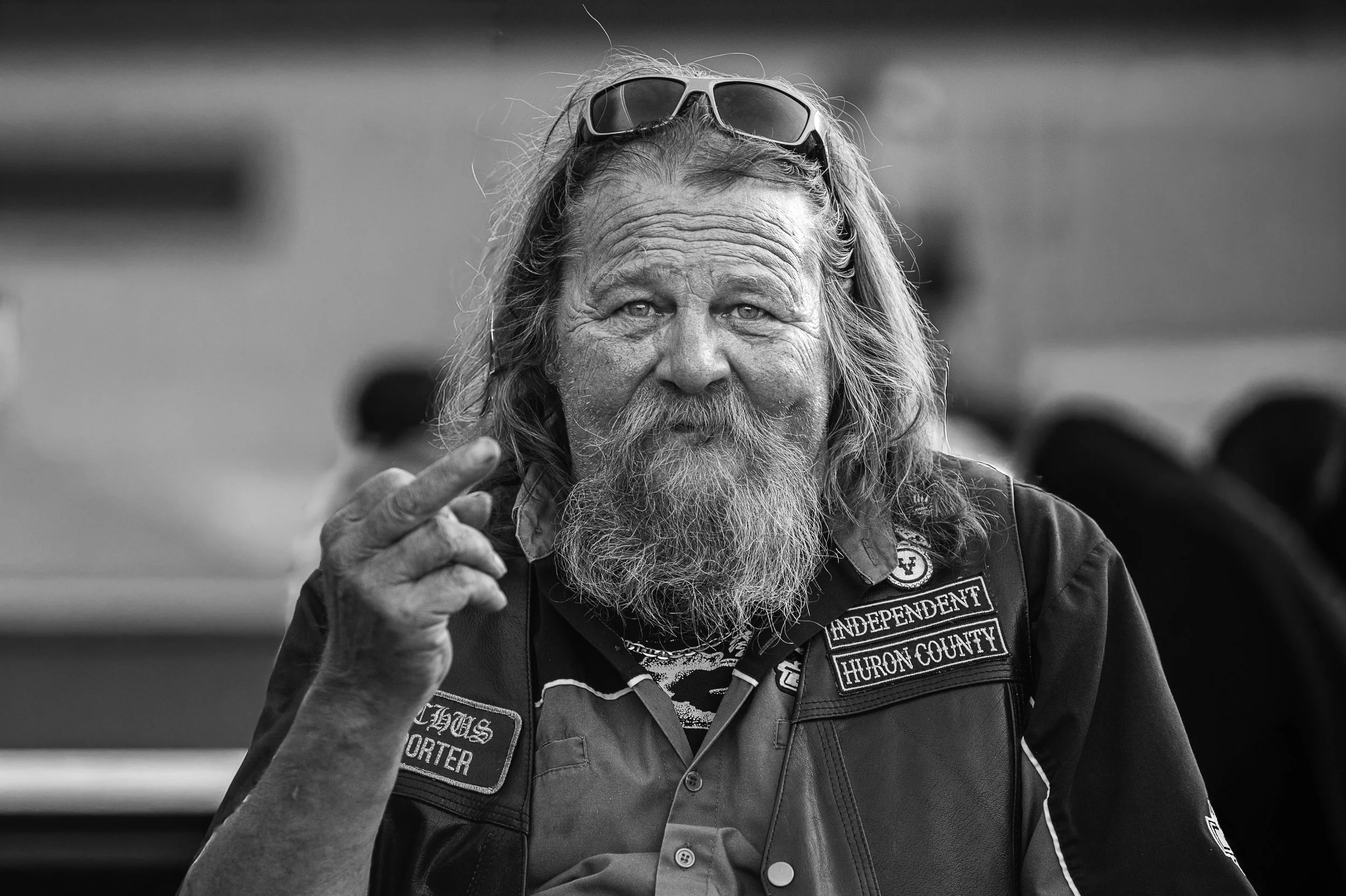 A bearded man with long hair, wearing sunglasses on his head,  dressed in a leather vest with patches, motorcycle.
