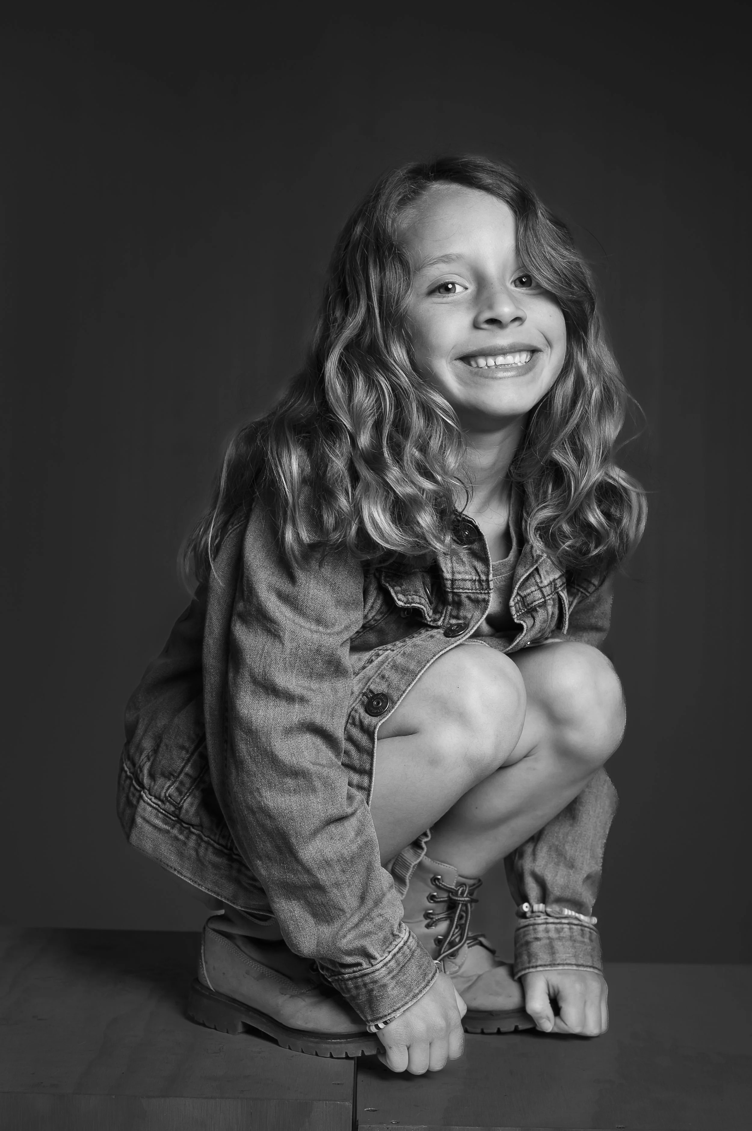Black and white photo of a smiling girl with long curly hair, wearing a denim jacket, shorts, and boots, squatting on a wooden surface against a dark background.