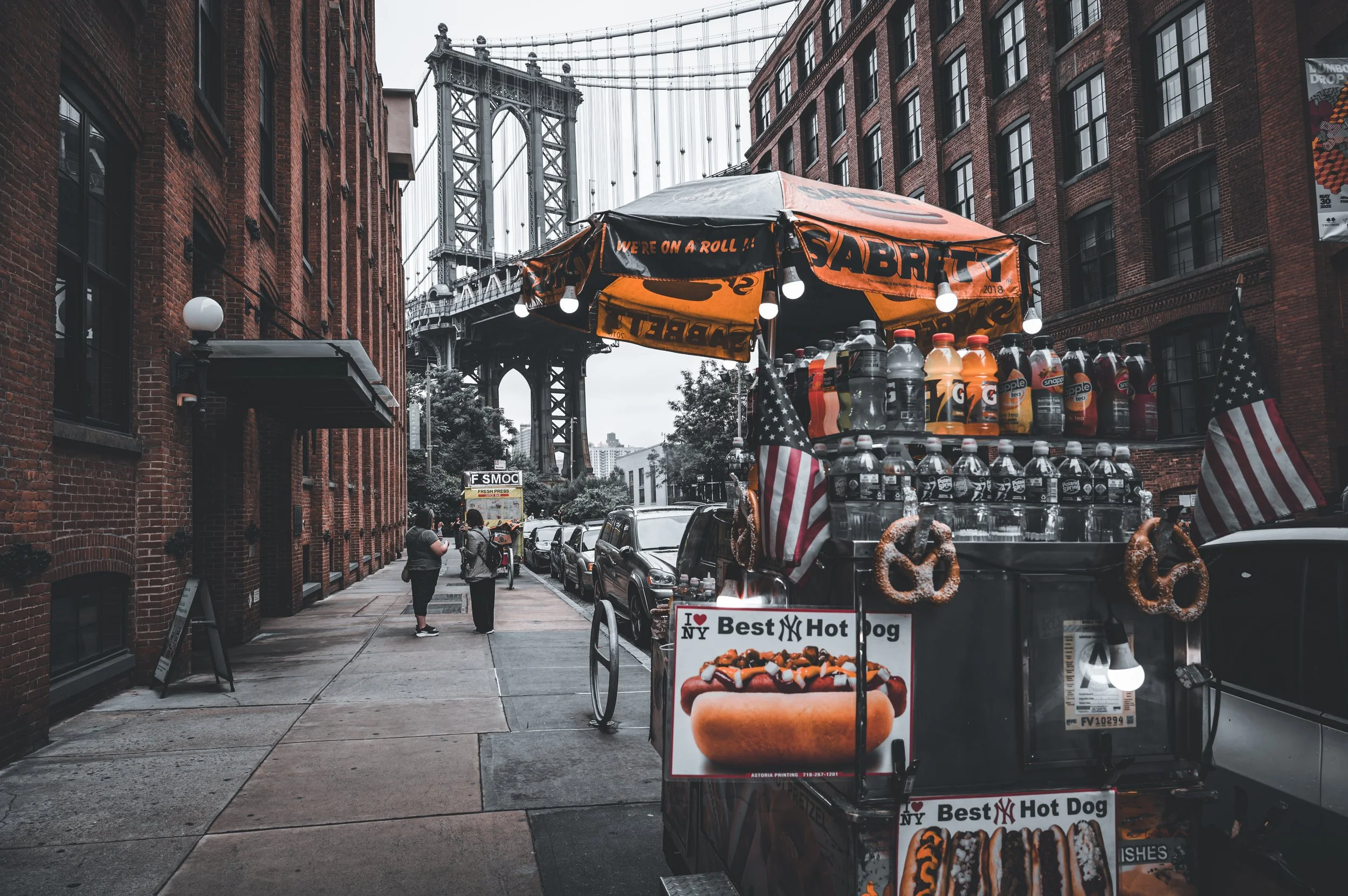 Street scene showing a hot dog stand decorated with American flags and pretzels, with the New York City Bridge in the background and pedestrians walking on the sidewalk.