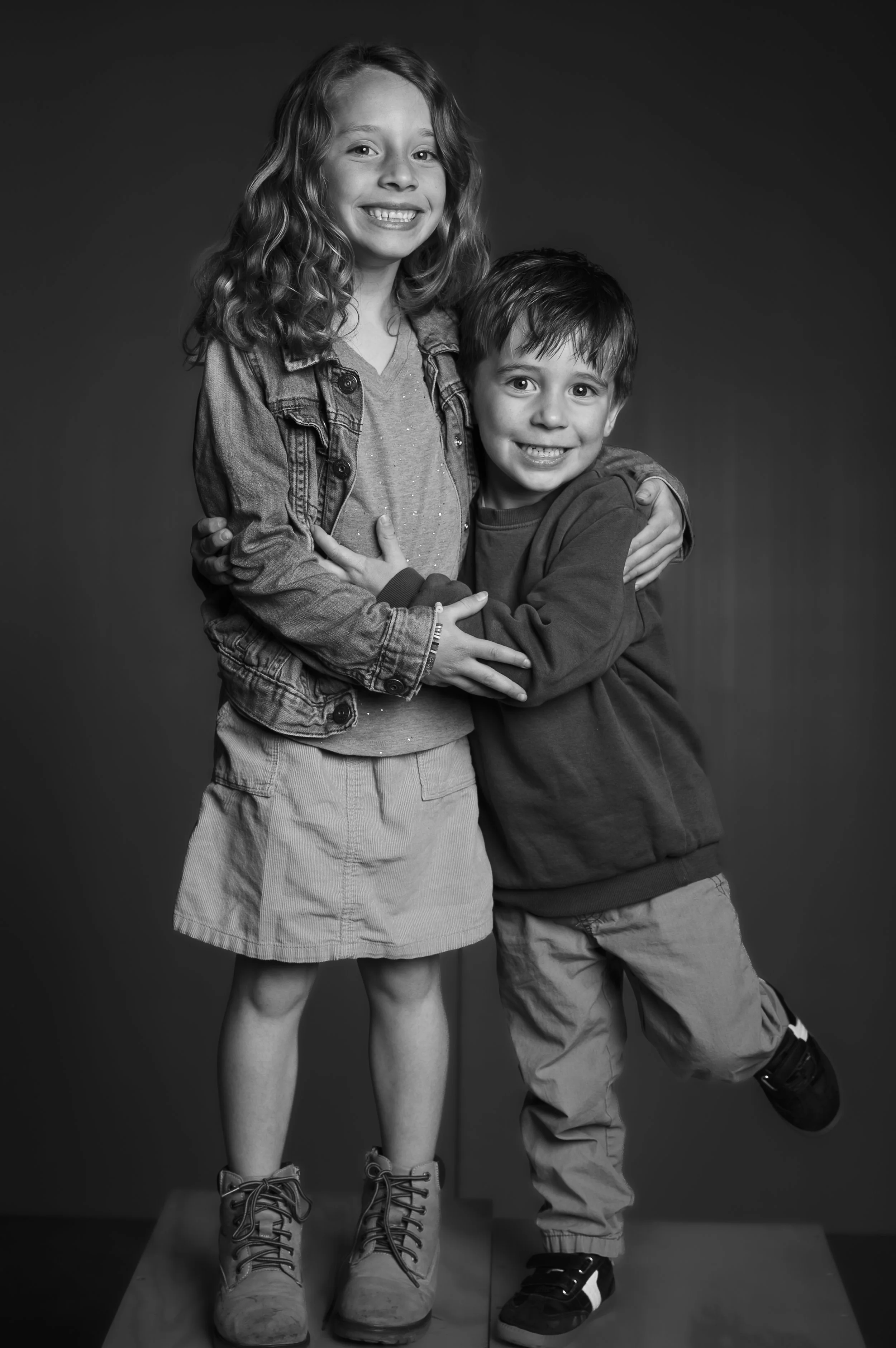 Black and white photo of a girl and a boy smiling and hugging against a plain background. The girl has wavy hair, wearing a denim jacket, a t-shirt, a skirt, and boots. The boy has straight hair, wearing a hoodie, loose pants, and sneakers.