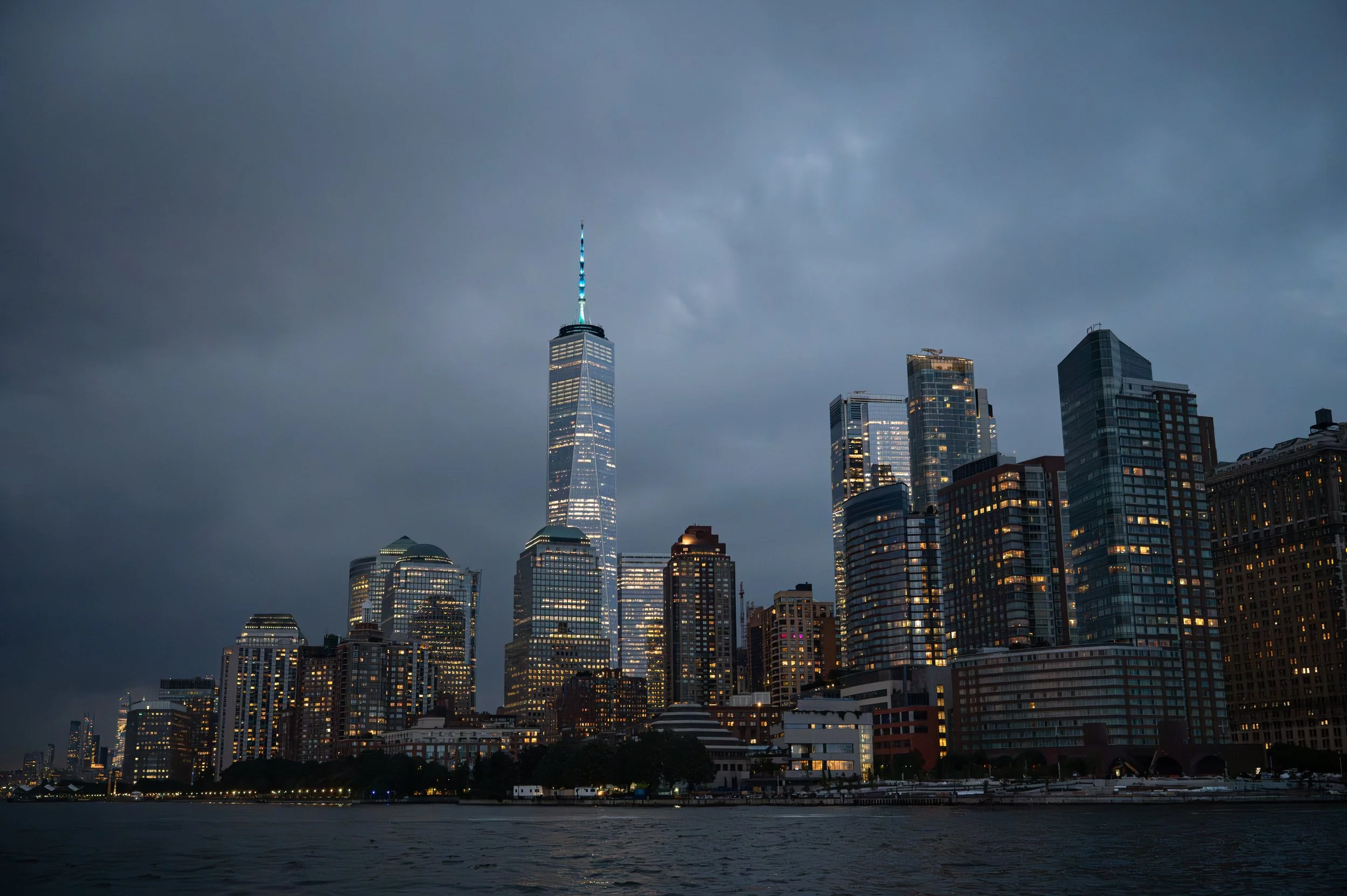 Nighttime view of Manhattan skyline with One World Trade Center and illuminated skyscrapers over the water.