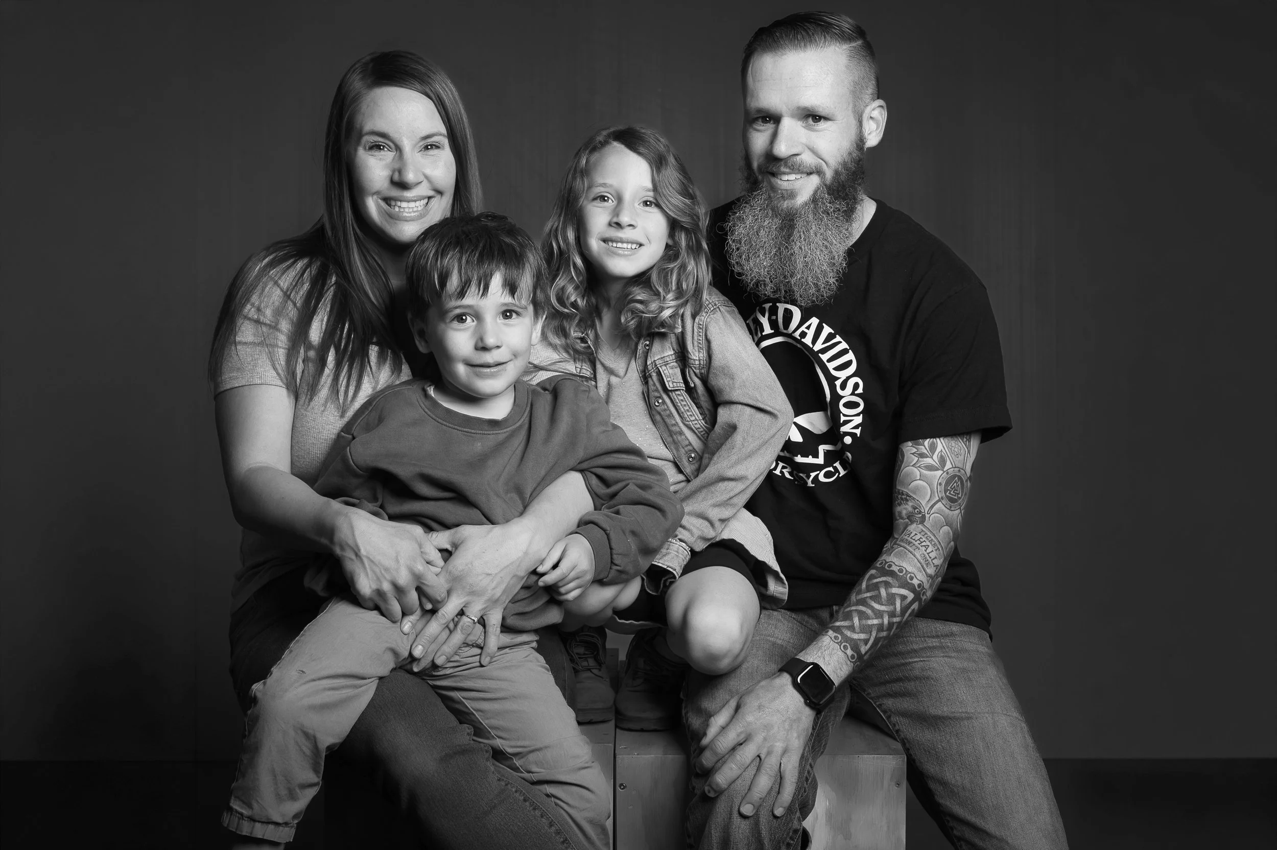 Black and white photo of a family of five sitting together, smiling. The family includes a woman, a man with a beard and tattoos, and three children, all sitting on and around a wooden box against a dark background.