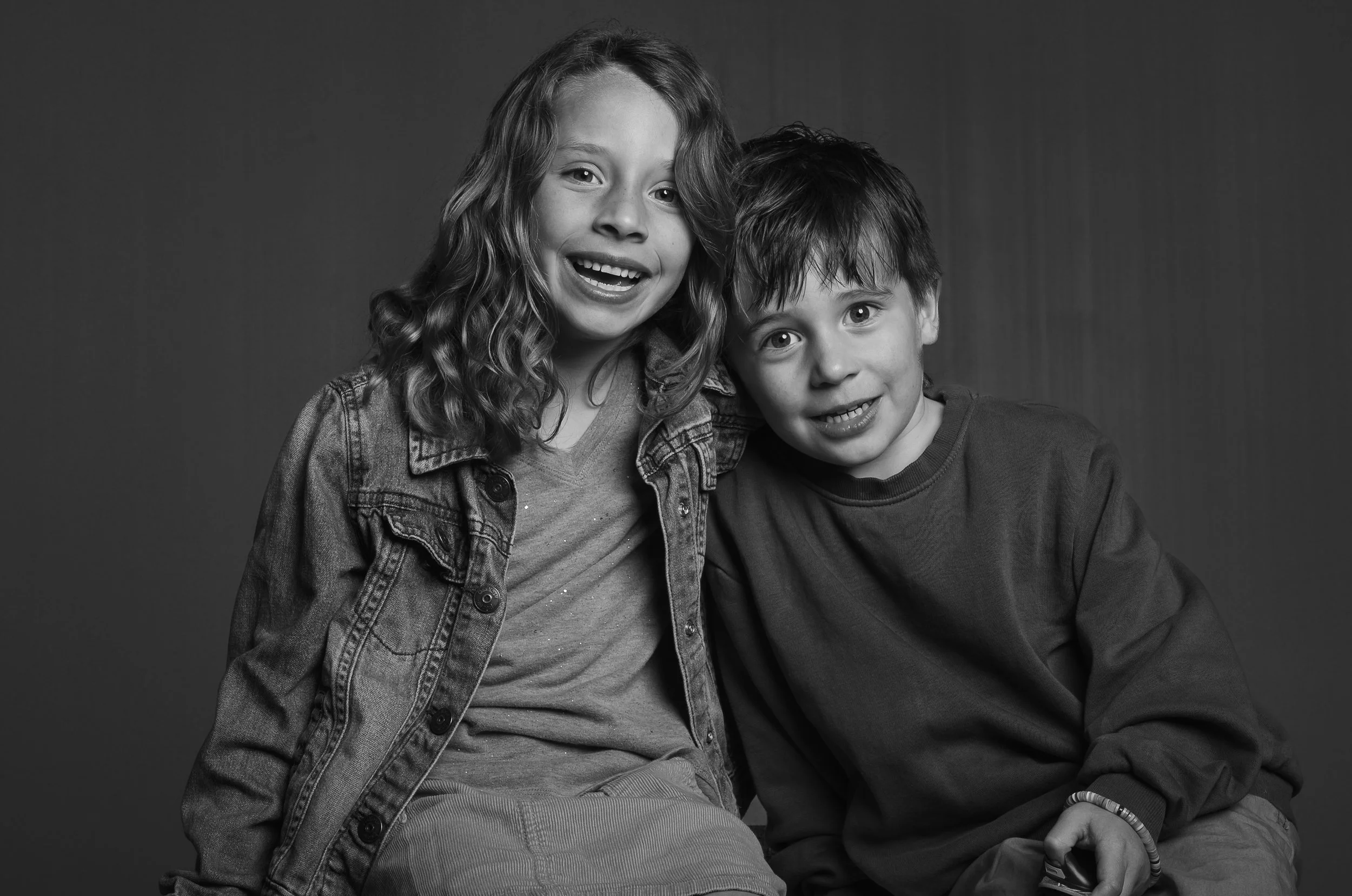 Black and white photo of a girl and boy smiling, seated close together, with the girl wearing a denim jacket and the boy in a long-sleeve shirt.