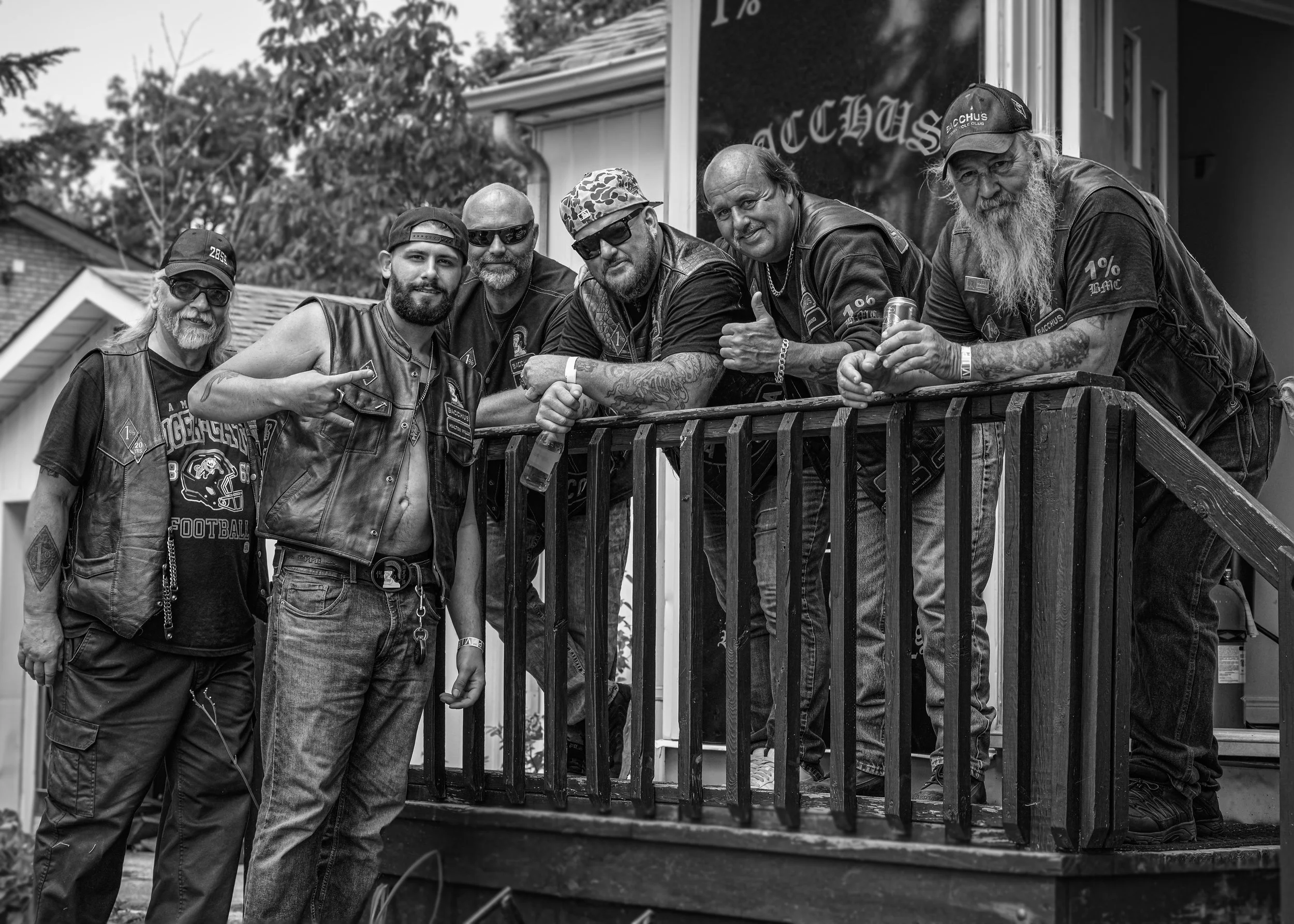 A group of seven men, mostly with beards and tattoos, standing on a porch railing. They are wearing biker vests or leather jackets, some with patches, and caps. One man is shirtless under his vest. They are smiling and holding drinks, with one giving