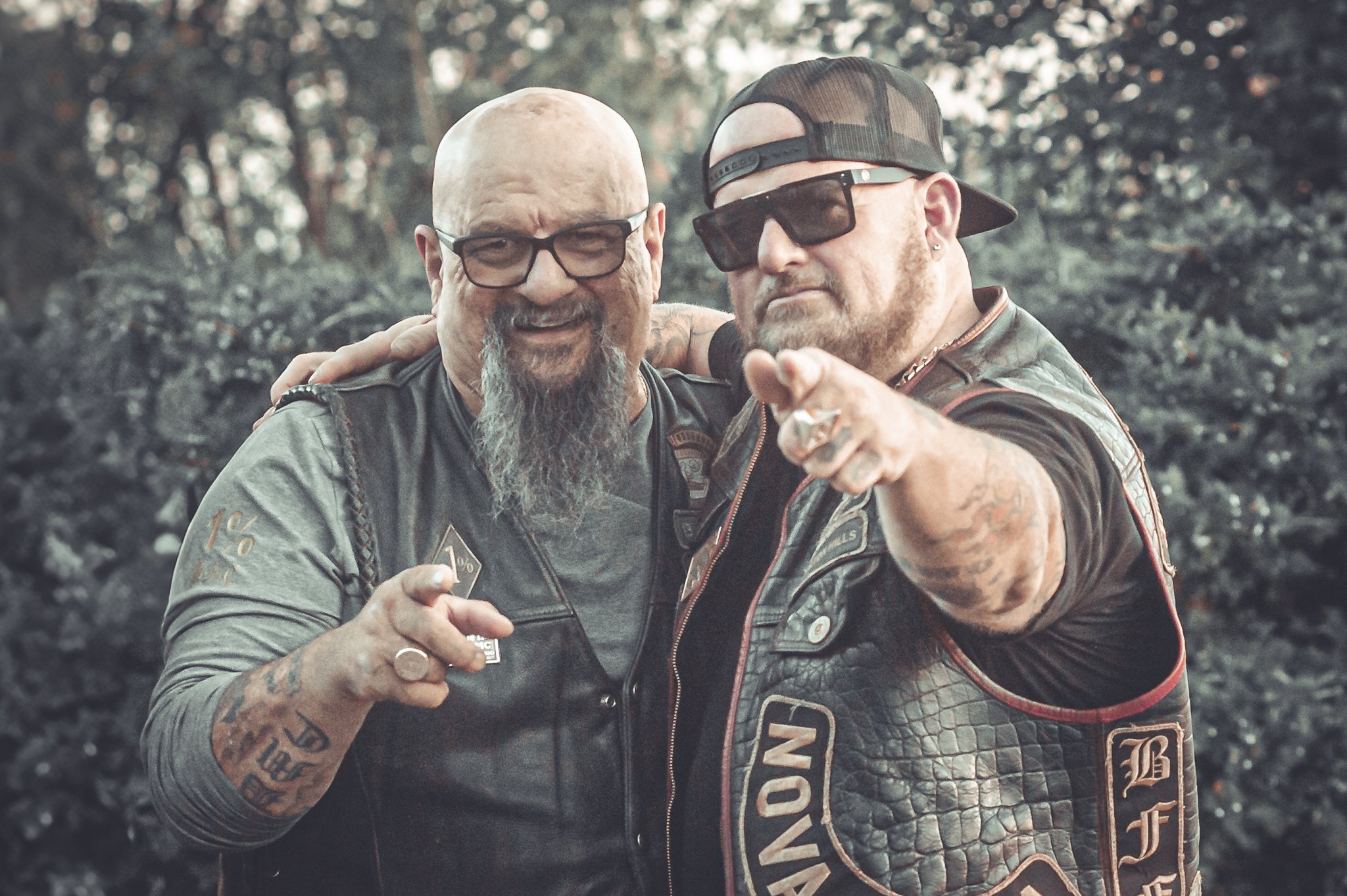 Two men wearing sunglasses and leather vests, one with a bald head and gray beard, the other with a baseball cap and a beard, standing outdoors with a background of trees. One man points at the camera.
