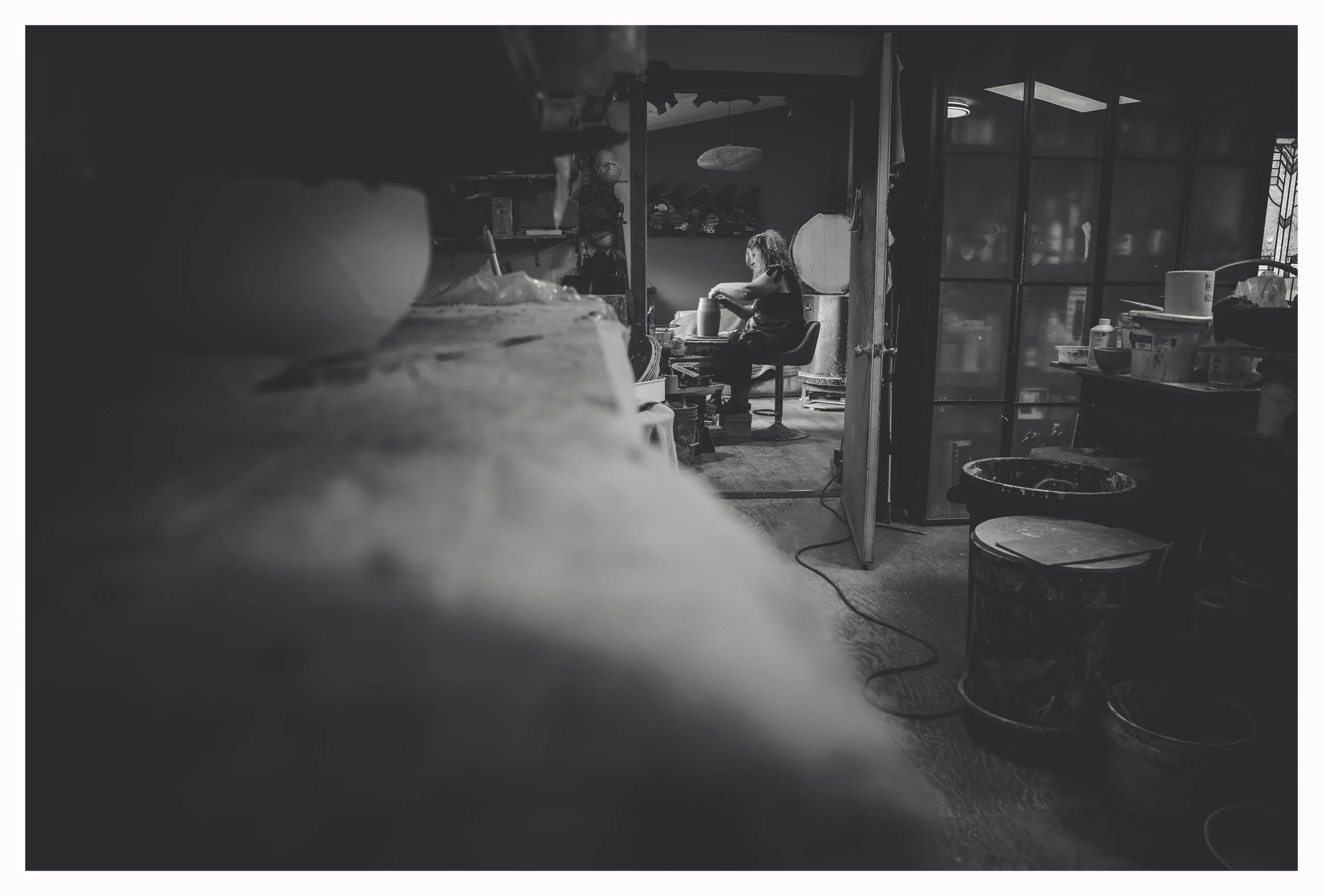 A woman working at a pottery wheel in a pottery studio, surrounded by shelves with pottery supplies, with a cluttered workspace and jars in the background.