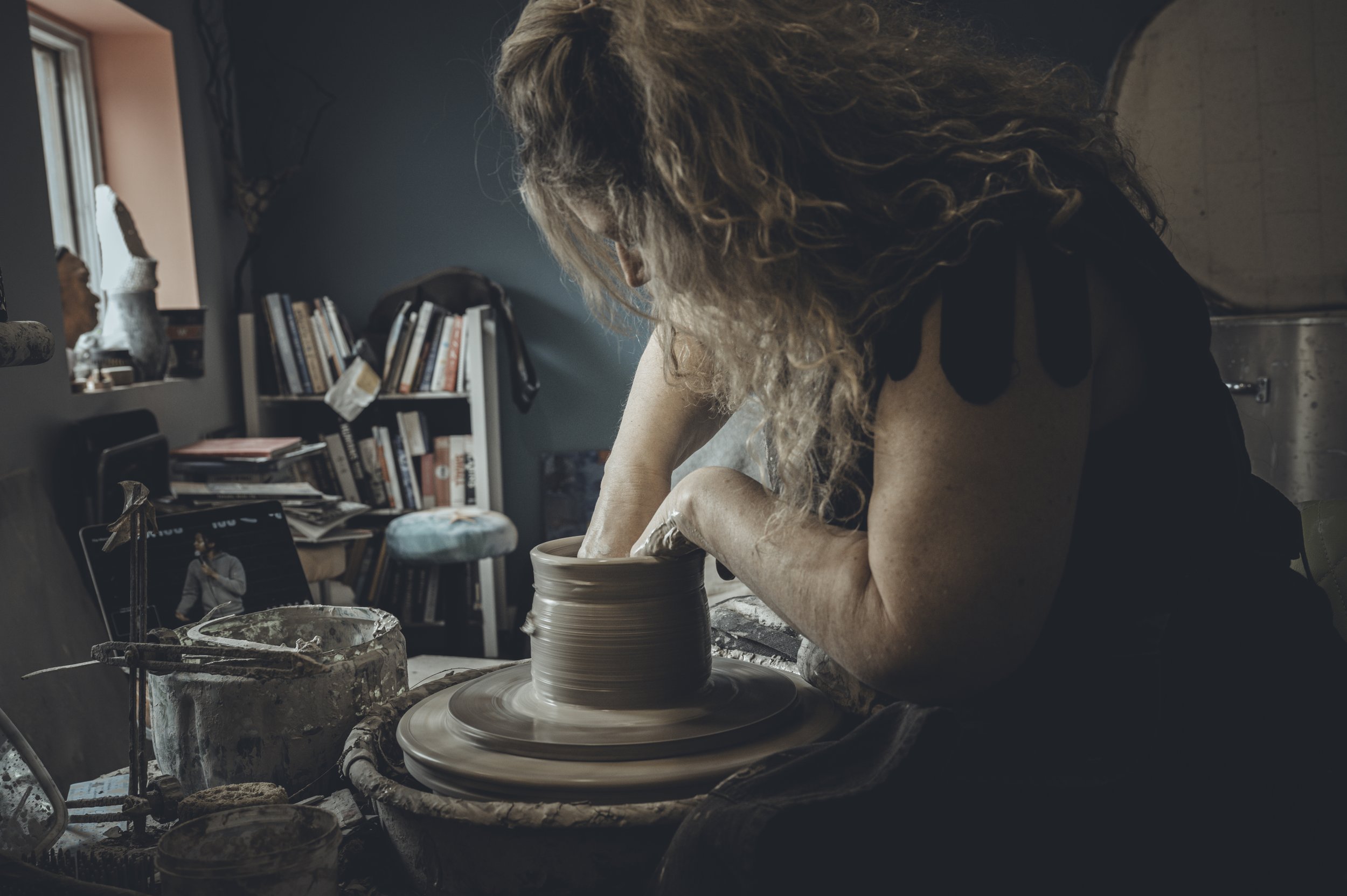 A woman creating pottery on a wheel in a craft studio, surrounded by art supplies and books.  Brand Photography Potter