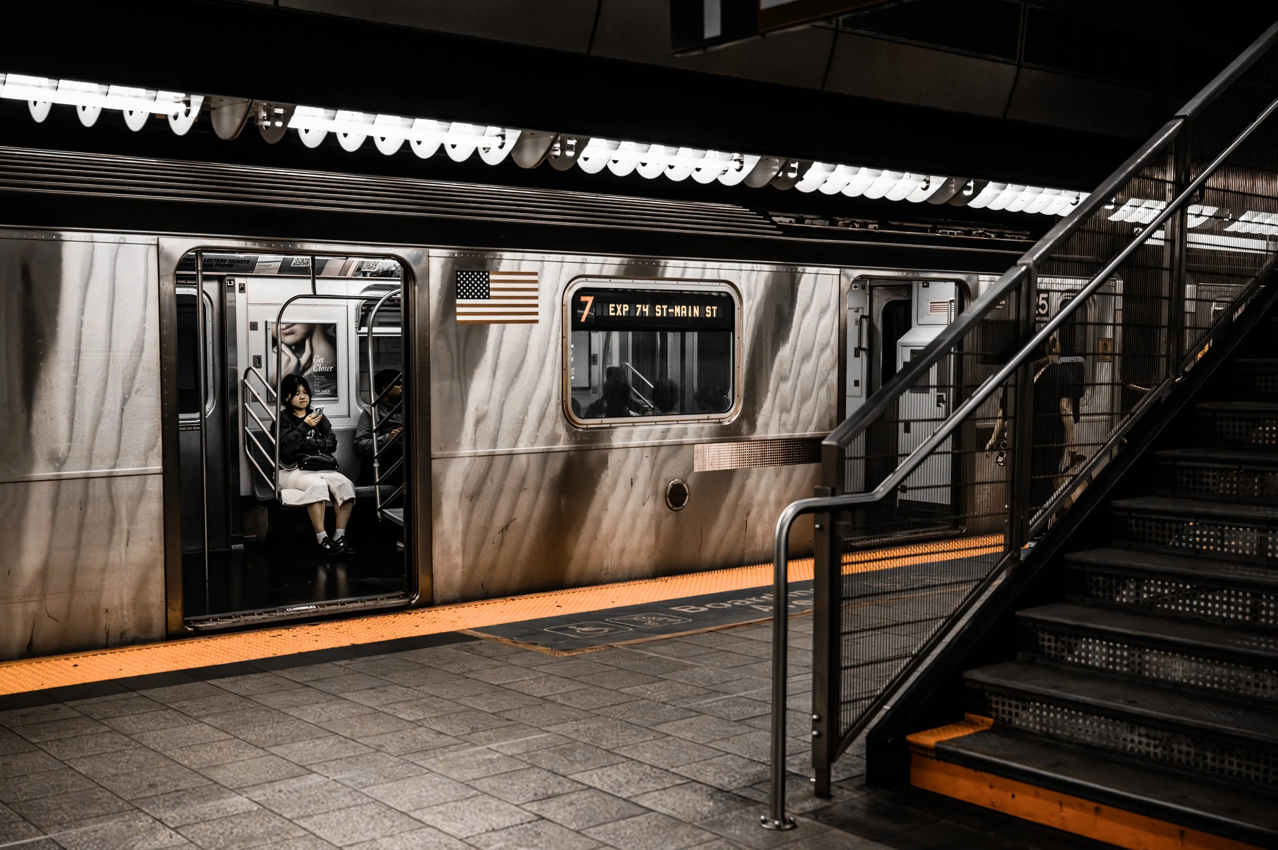 A subway train at an underground station with an open door showing two passengers sitting inside. One passenger is a woman wearing a black jacket and white skirt, looking at her phone. A stairway on the right leads to an upper platform. New York City