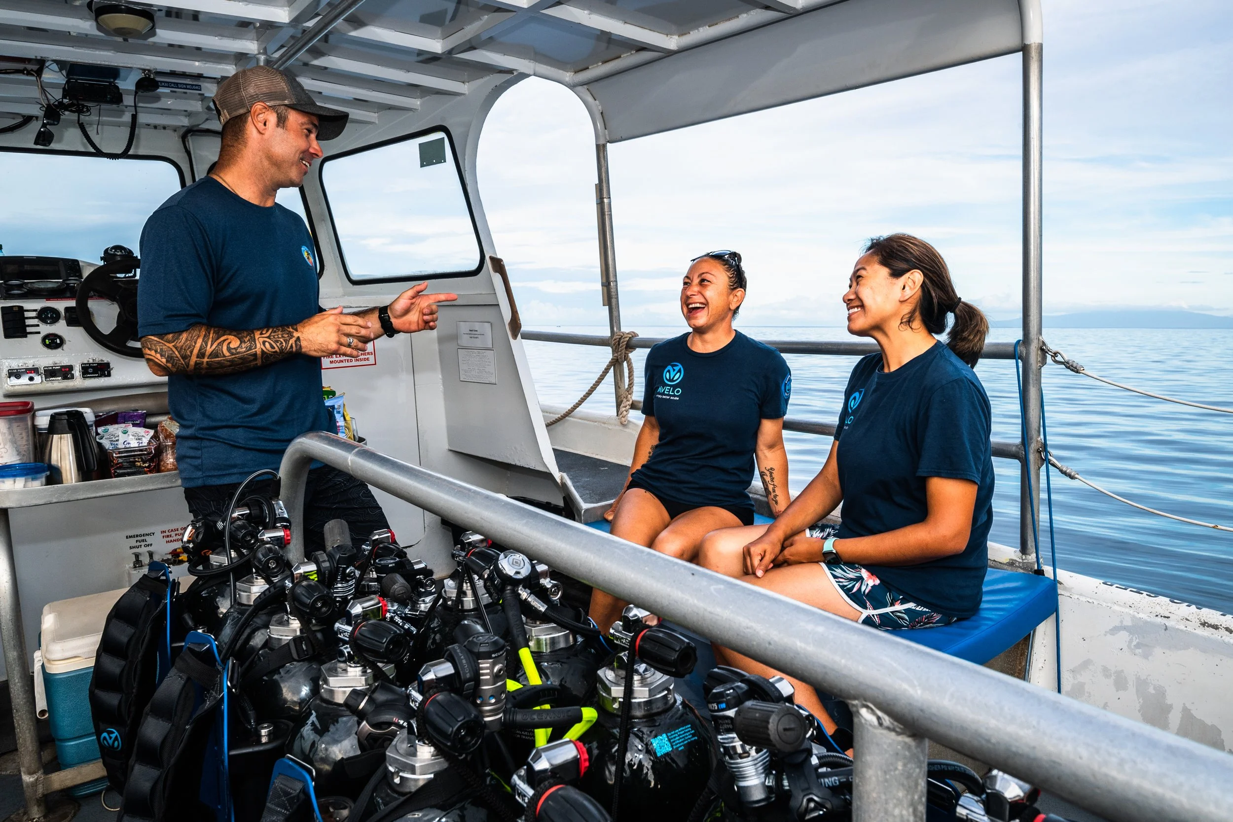 Two women and a man on a boat having a conversation, with scuba tanks in the foreground and open water in the background.