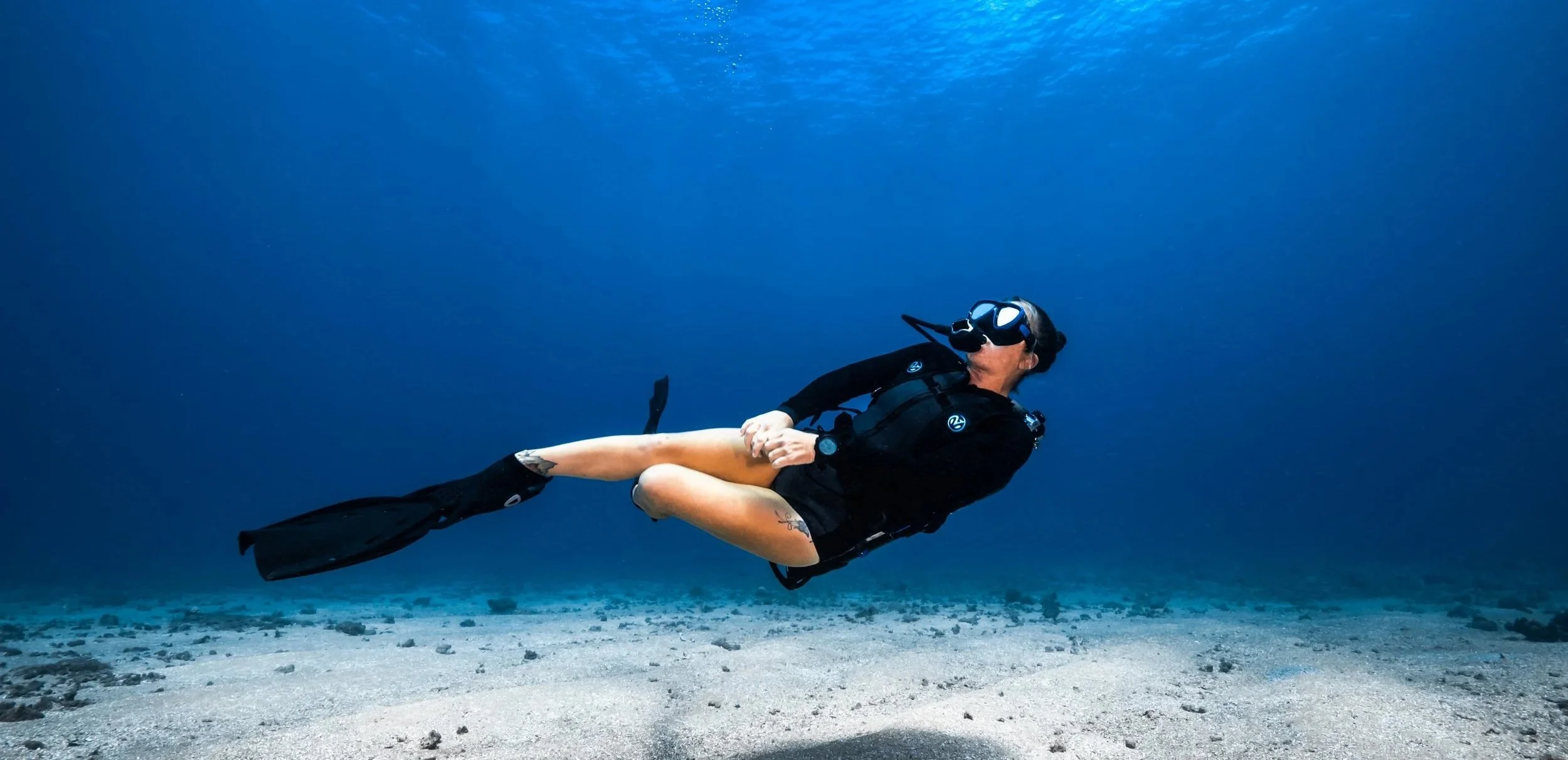 Person scuba diving underwater with mask and fins, swimming above sandy ocean floor with rocks.