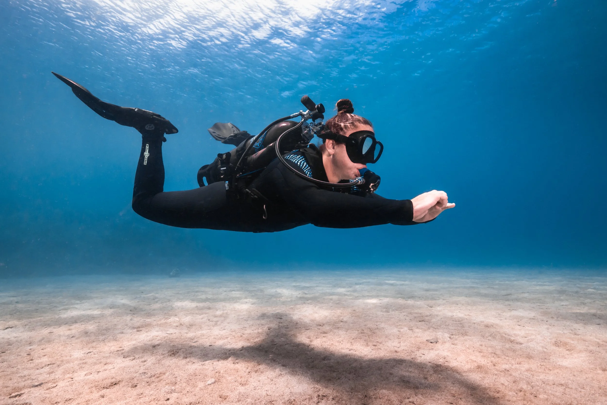A woman scuba diving underwater, wearing a black wetsuit, scuba mask, and fins, swimming over a sandy ocean floor with clear blue water above.