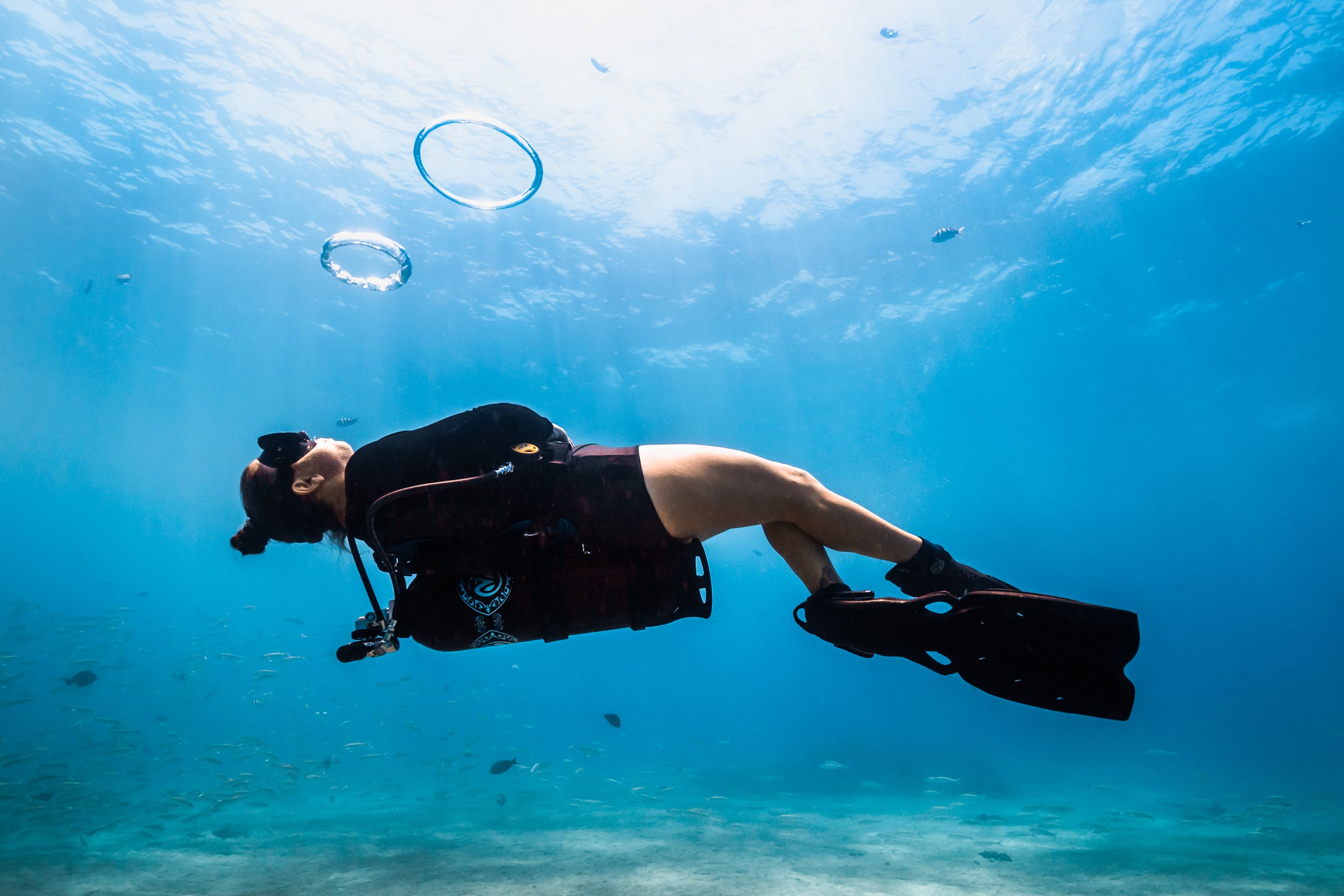 A woman in scuba gear swimming underwater with a clear blue ocean background, bubbles rising, and small fish swimming nearby.