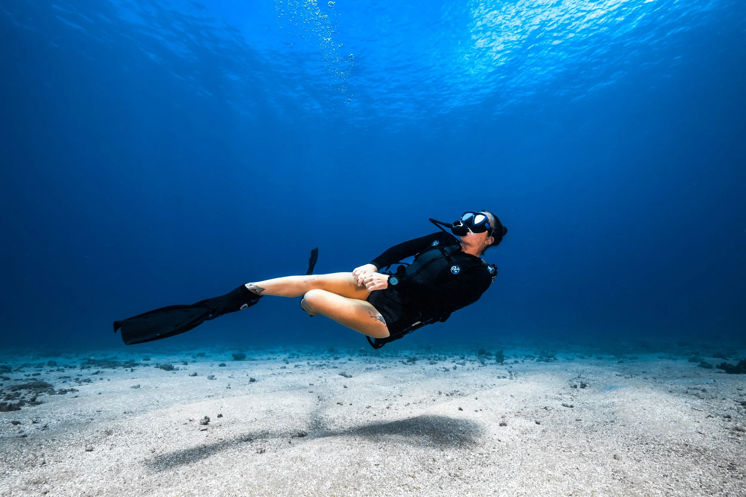 Person in scuba gear floating underwater above sandy ocean floor with deep blue water surrounding.