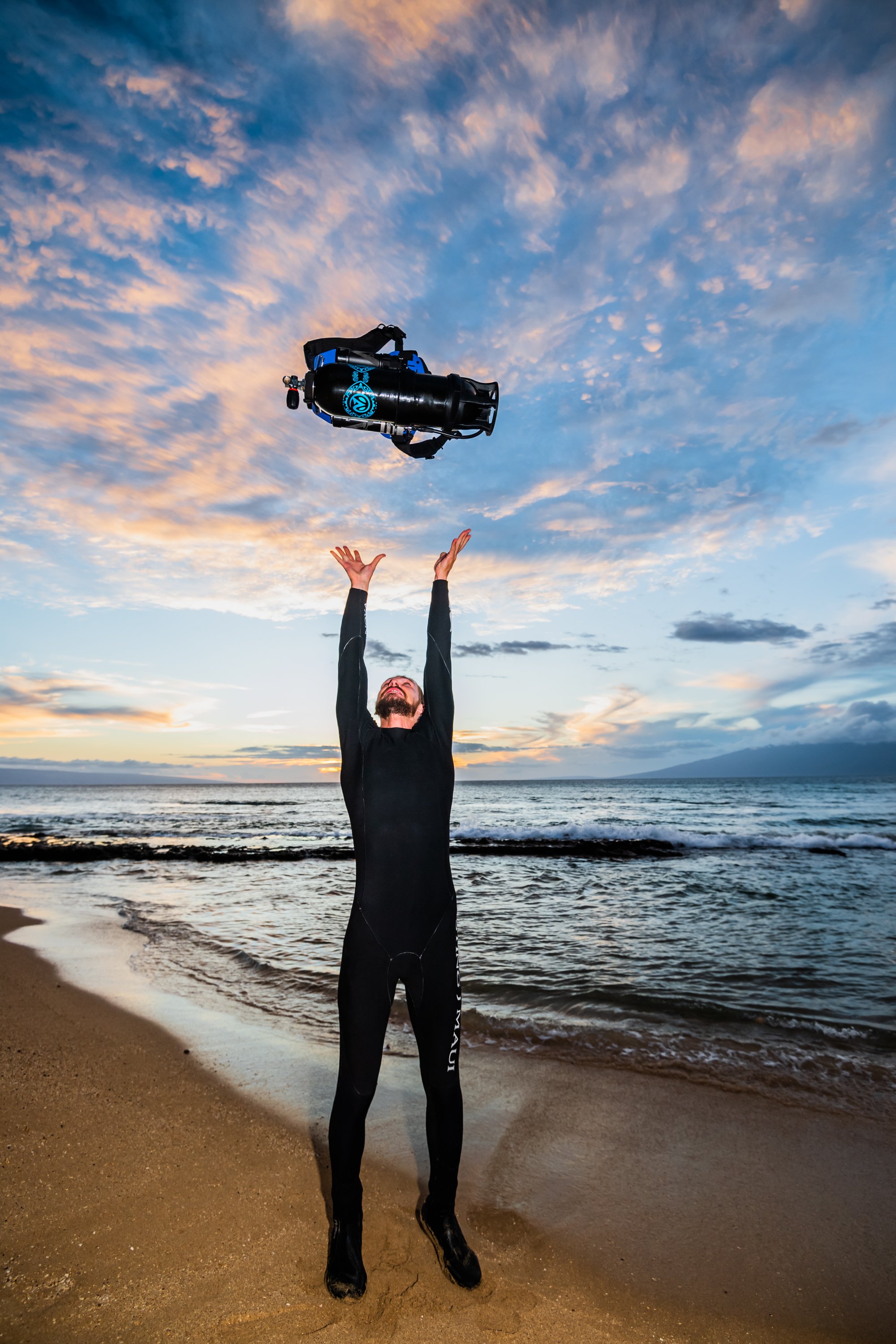 A man in a wetsuit on the beach tossing a scuba tank into the air during sunset.