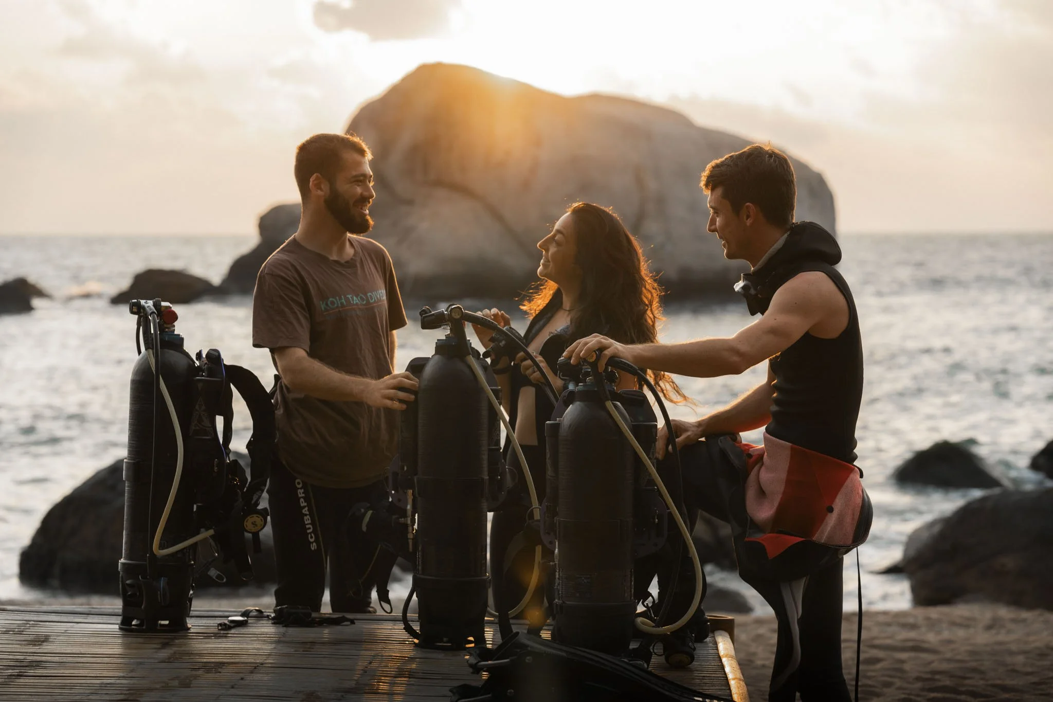 Three scuba divers preparing their gear on a dock at sunset near the ocean, with a large rock formation in the background.