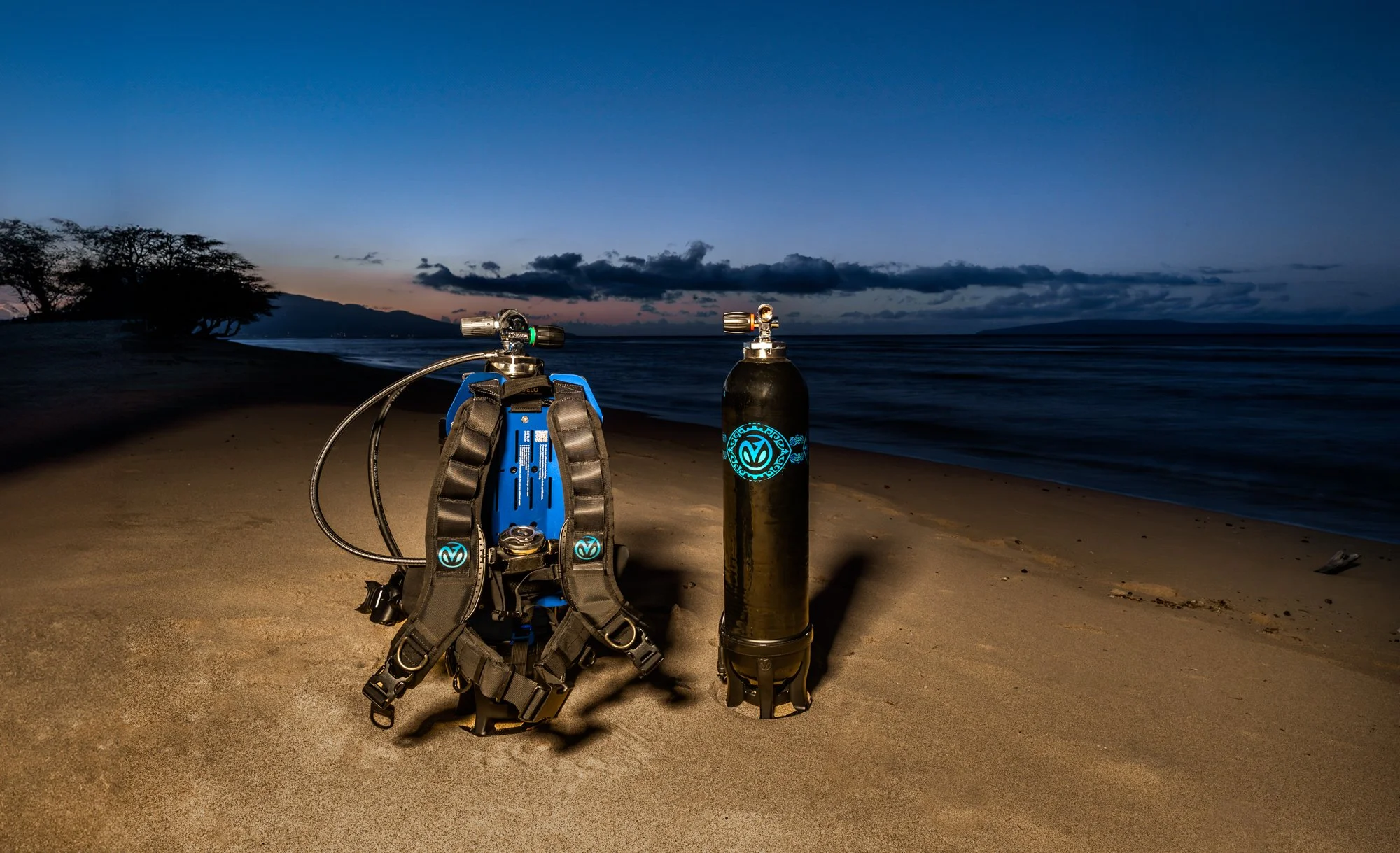 Open scuba tank and backpack on sandy beach at sunset with trees and ocean in the background.