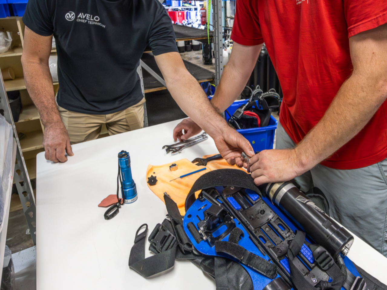 A woman scuba diving underwater, wearing a black wetsuit, scuba mask, and fins, swimming over a sandy ocean floor with clear blue water above.