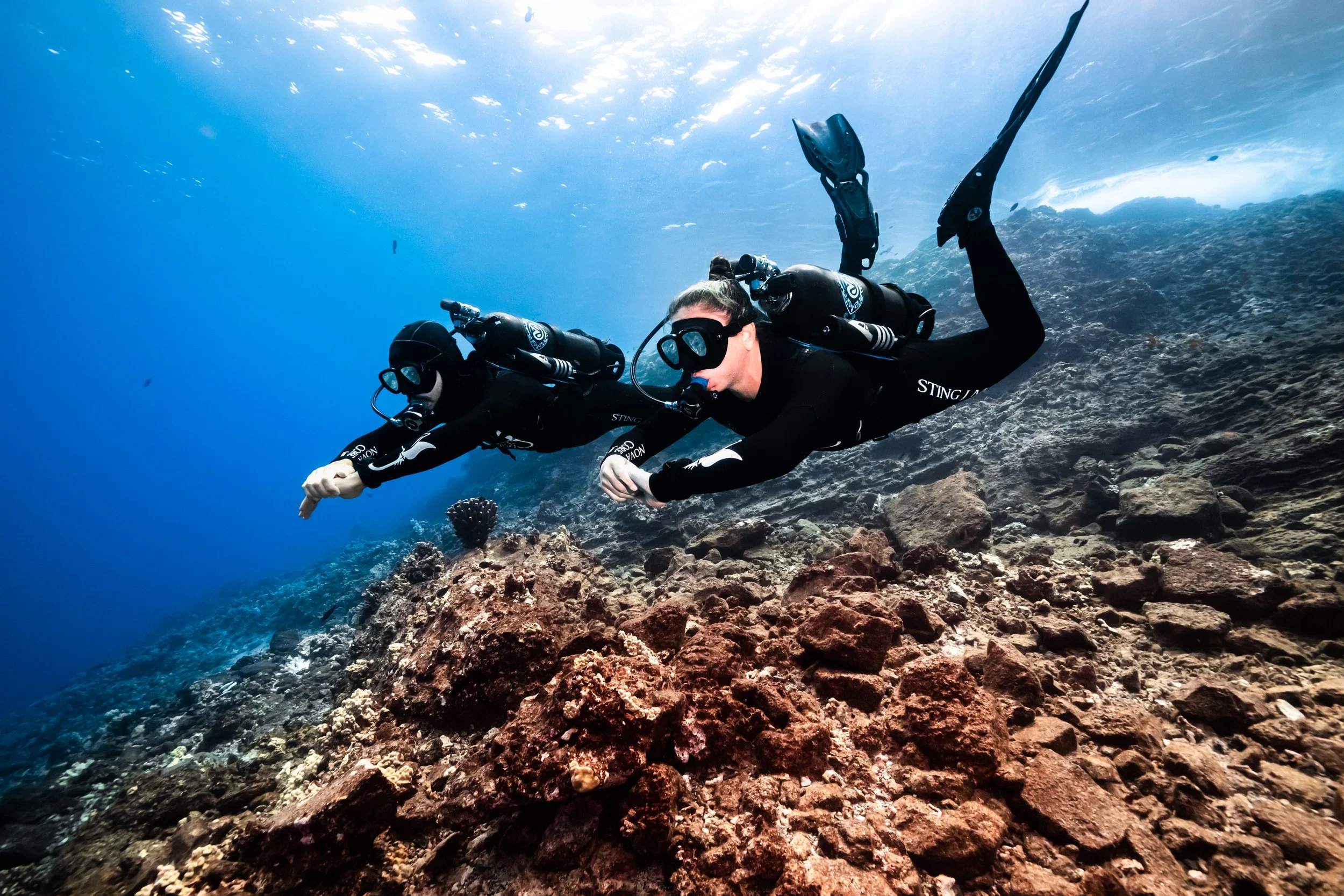 Two scuba divers in black wetsuits swimming underwater over a rocky coral reef.