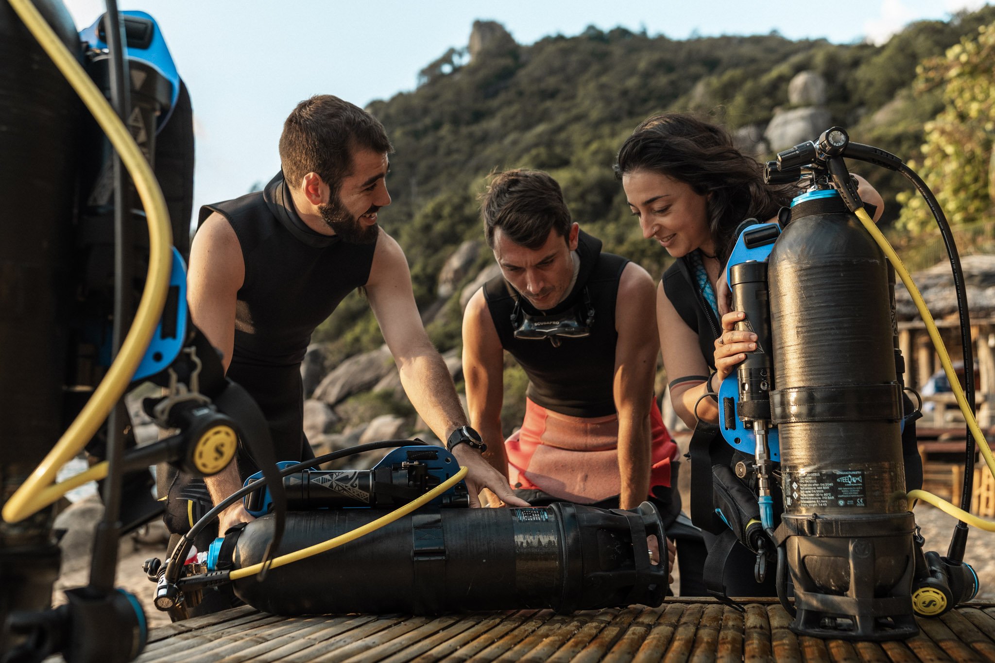 Three divers prepare their scuba gear outdoors on a wooden surface, with a natural landscape in the background.