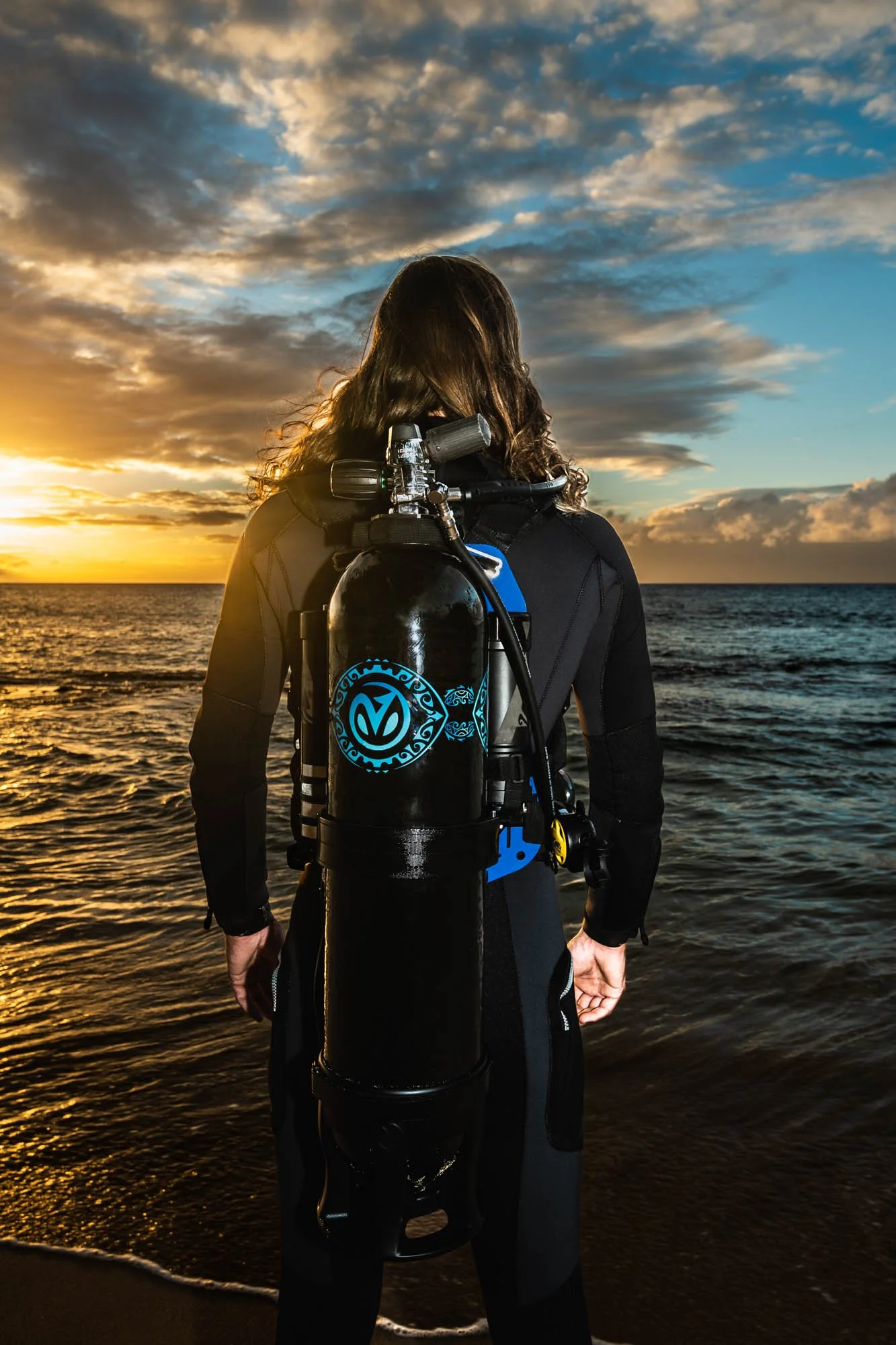 Person in wetsuit standing by the ocean at sunset, carrying a scuba tank on their back.