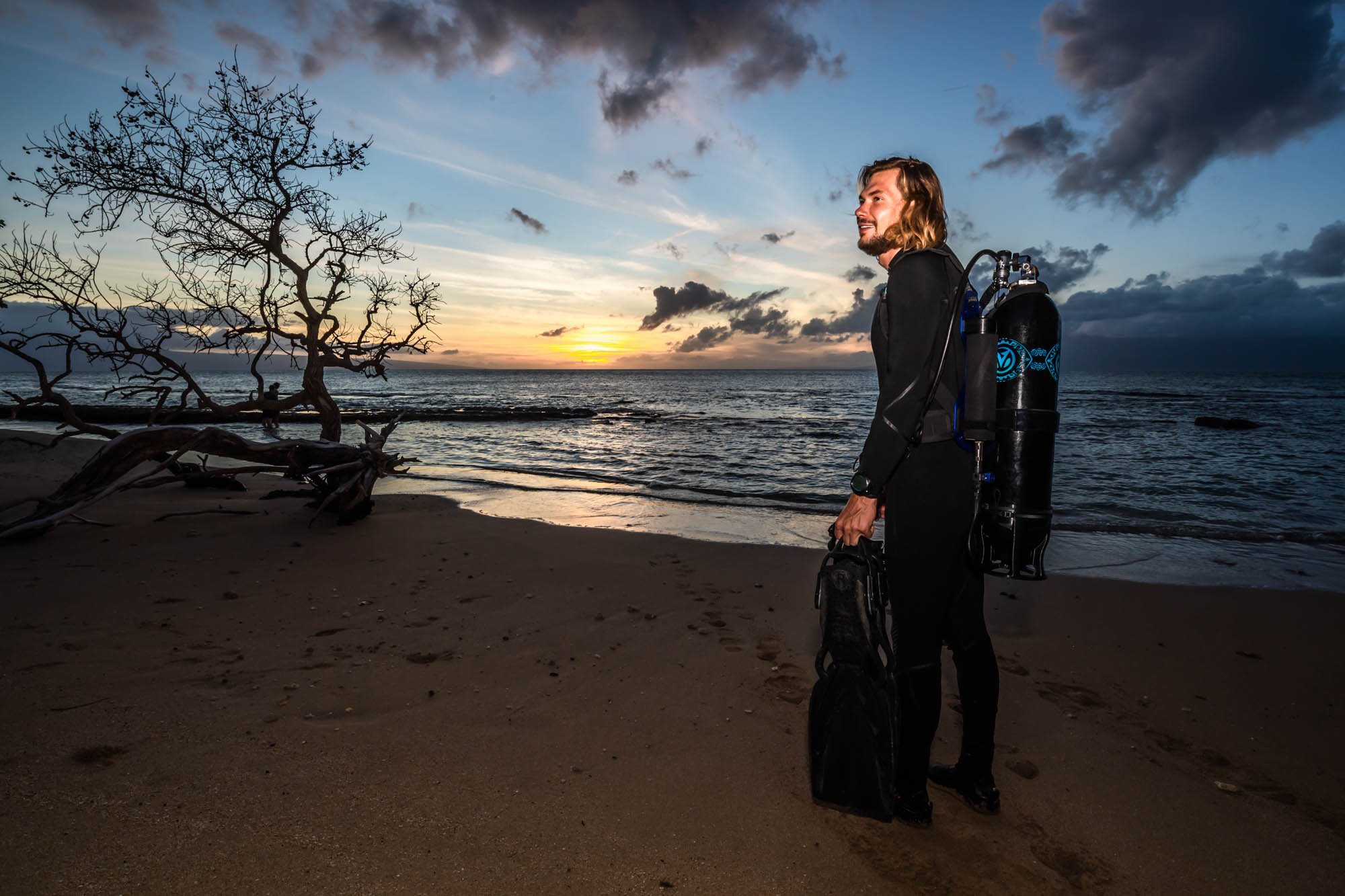 A man standing on a beach at sunset, wearing a wetsuit with scuba tanks on his back and holding a diving bag.