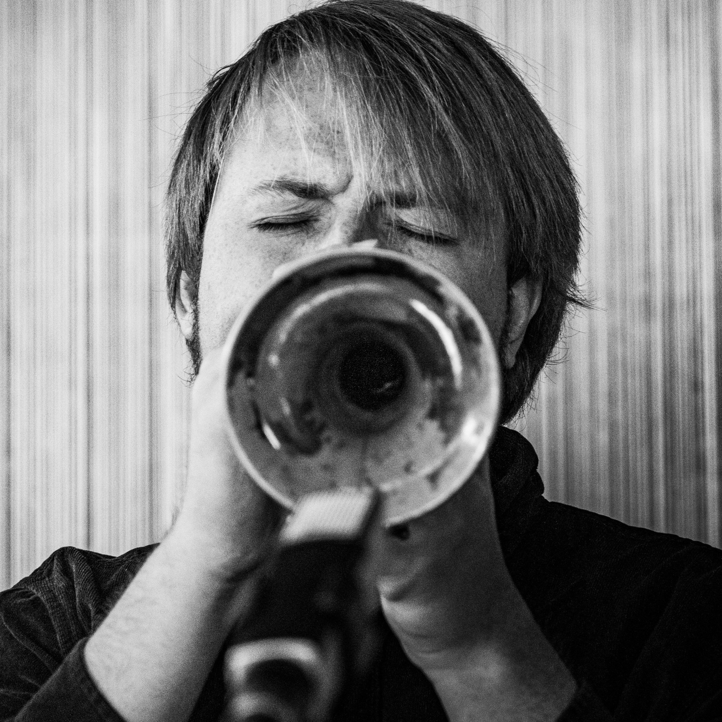 A black-and-white photo of a man with closed eyes aiming a trumpet directly at the camera.