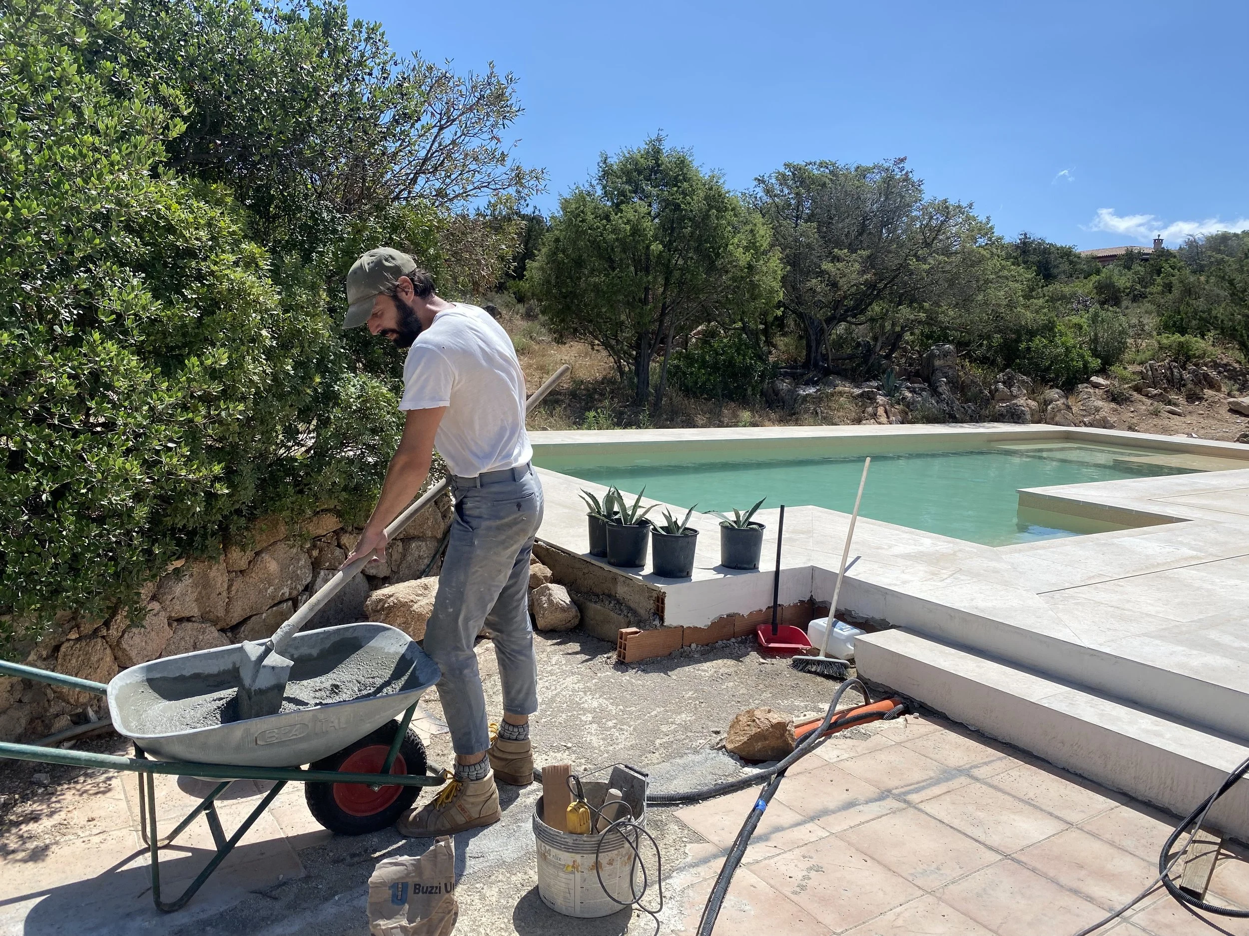 A man working on a construction site near a swimming pool, handling a wheelbarrow filled with cement, with trees, rocks, and a clear blue sky in the background.