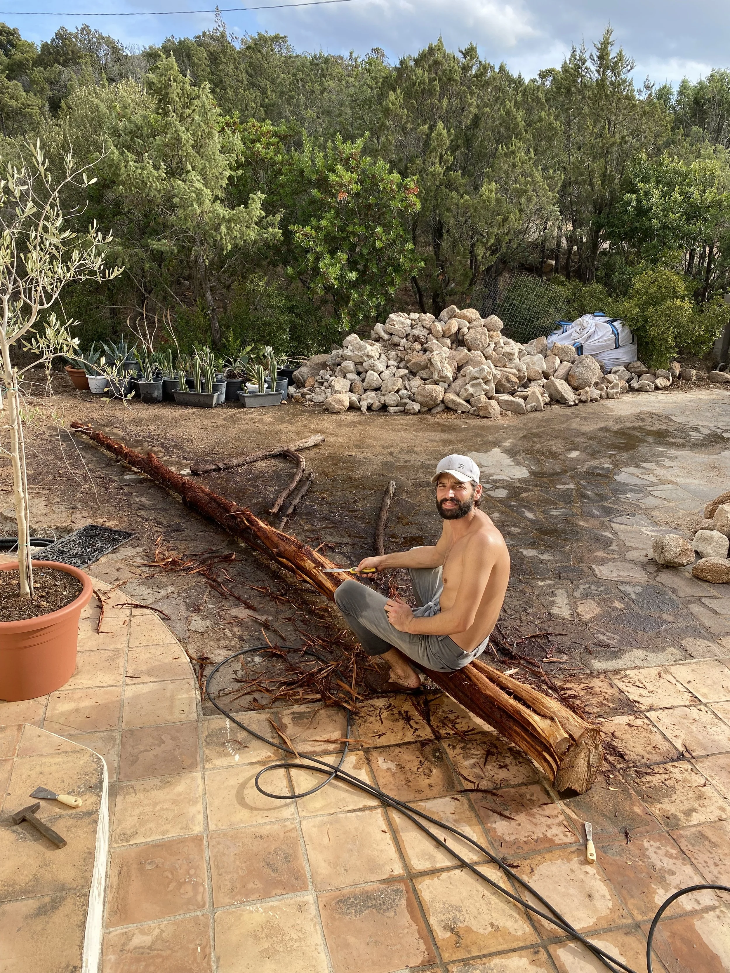 A shirtless man wearing a white cap is sitting on a fallen tree trunk, using a saw to cut it. The background features a garden with potted plants, rocks, and trees under a partly cloudy sky.