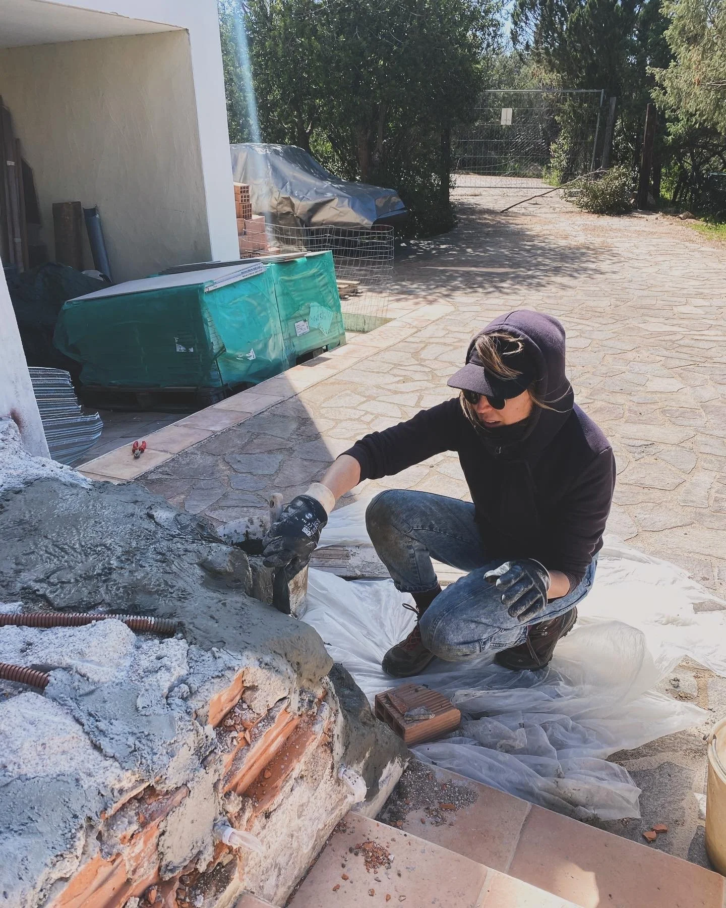 A person wearing sunglasses, a hoodie, and gloves, kneeling on the ground, working on a construction or repair project outdoors near a brick structure.