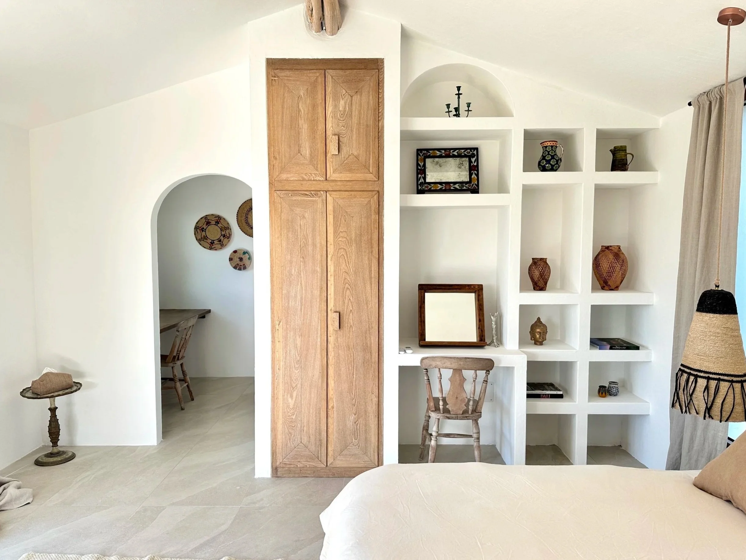 A bedroom with light-colored walls and tiled flooring. There is a built-in white bookshelf with decorative items and a small desk with a chair. A wooden closet door is centered in the room, and a small table with a hat on it stands near the closet. A
