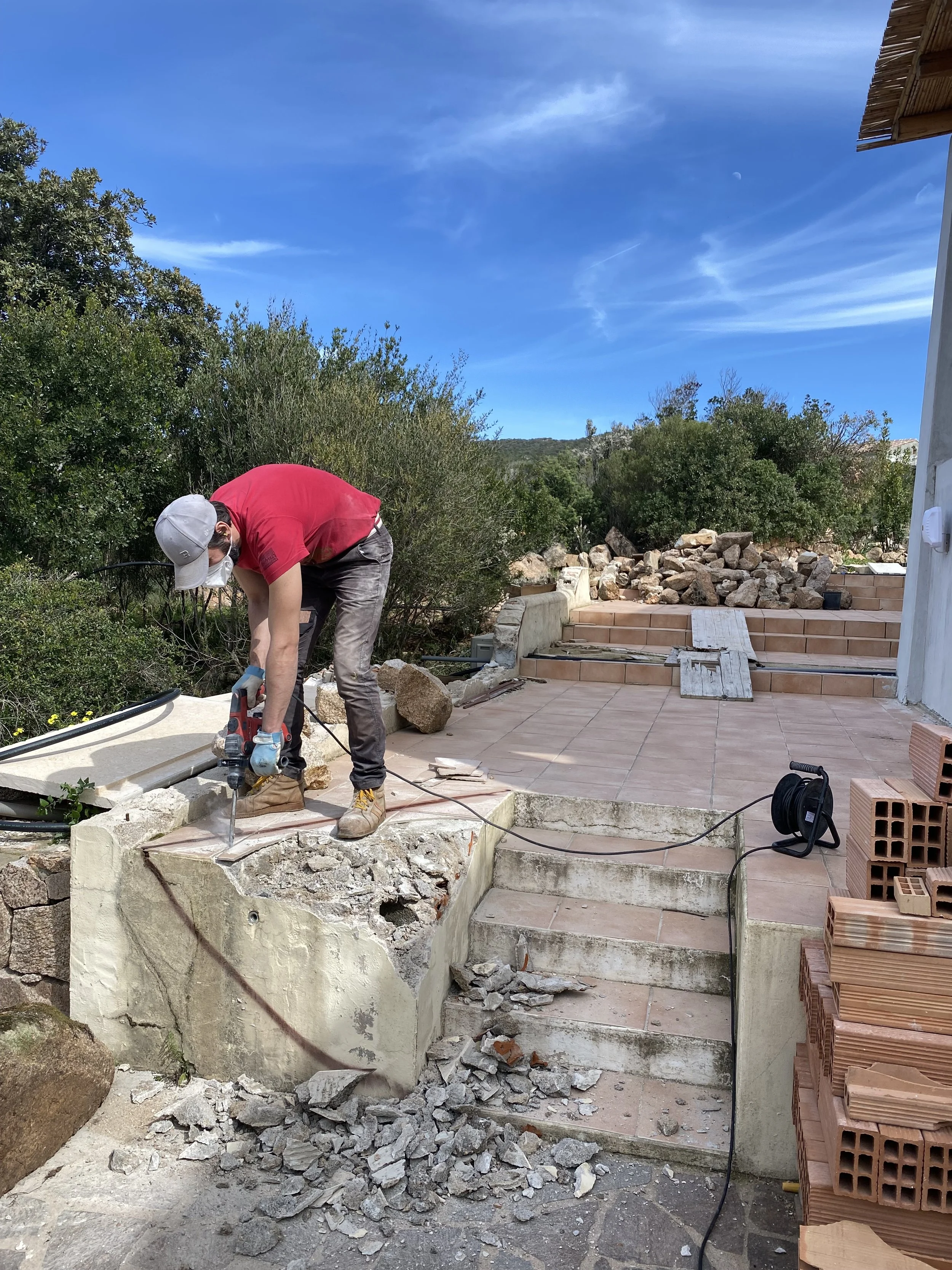 A person cutting concrete on a damaged section of a stairway outdoors, surrounded by construction materials and tools, with trees and a blue sky in the background.
