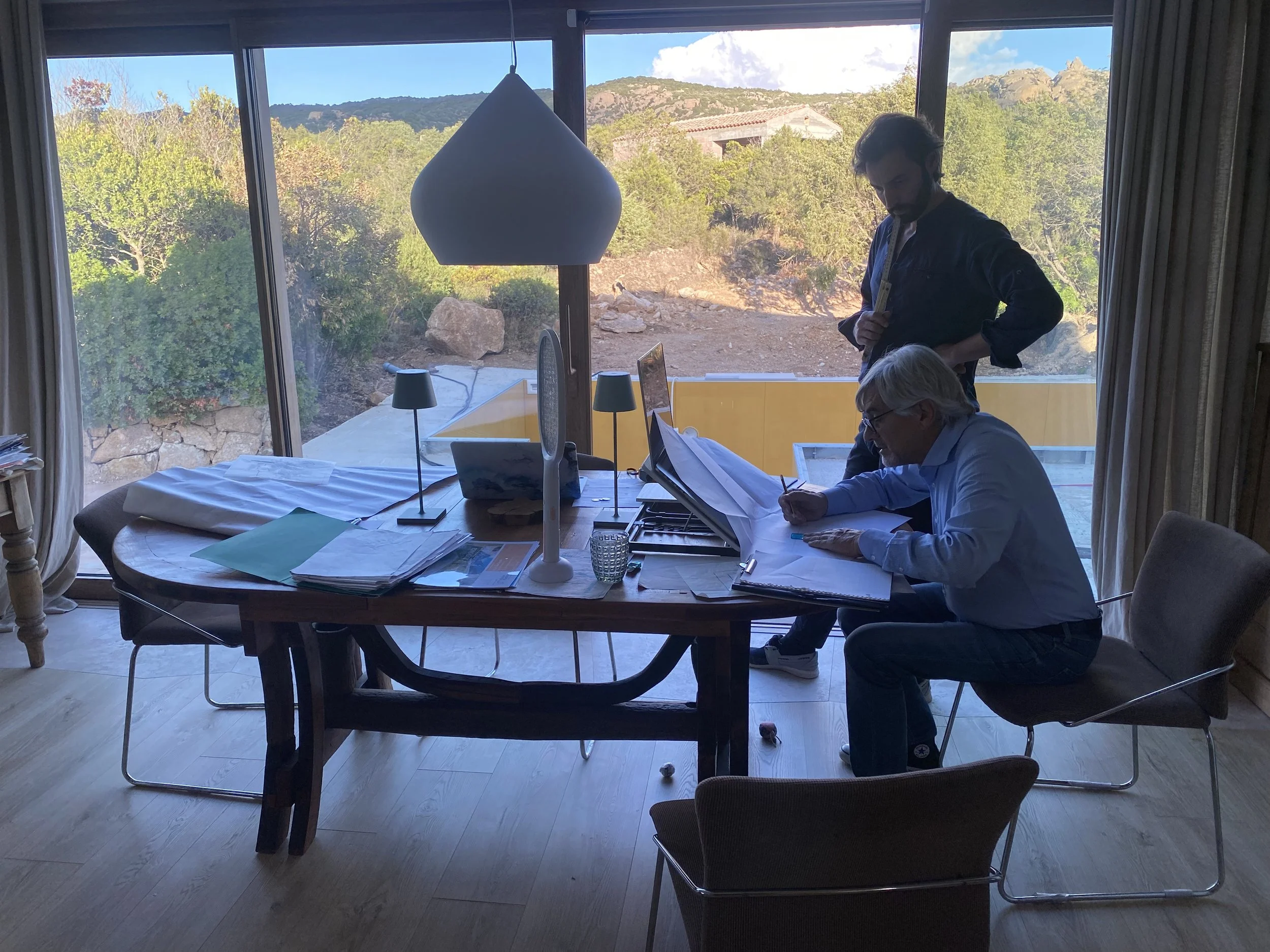 Two men working at a large desk in front of a window overlooking a rocky outdoor terrain with trees and mountains in the background. The desk is cluttered with papers, a computer, and office supplies.