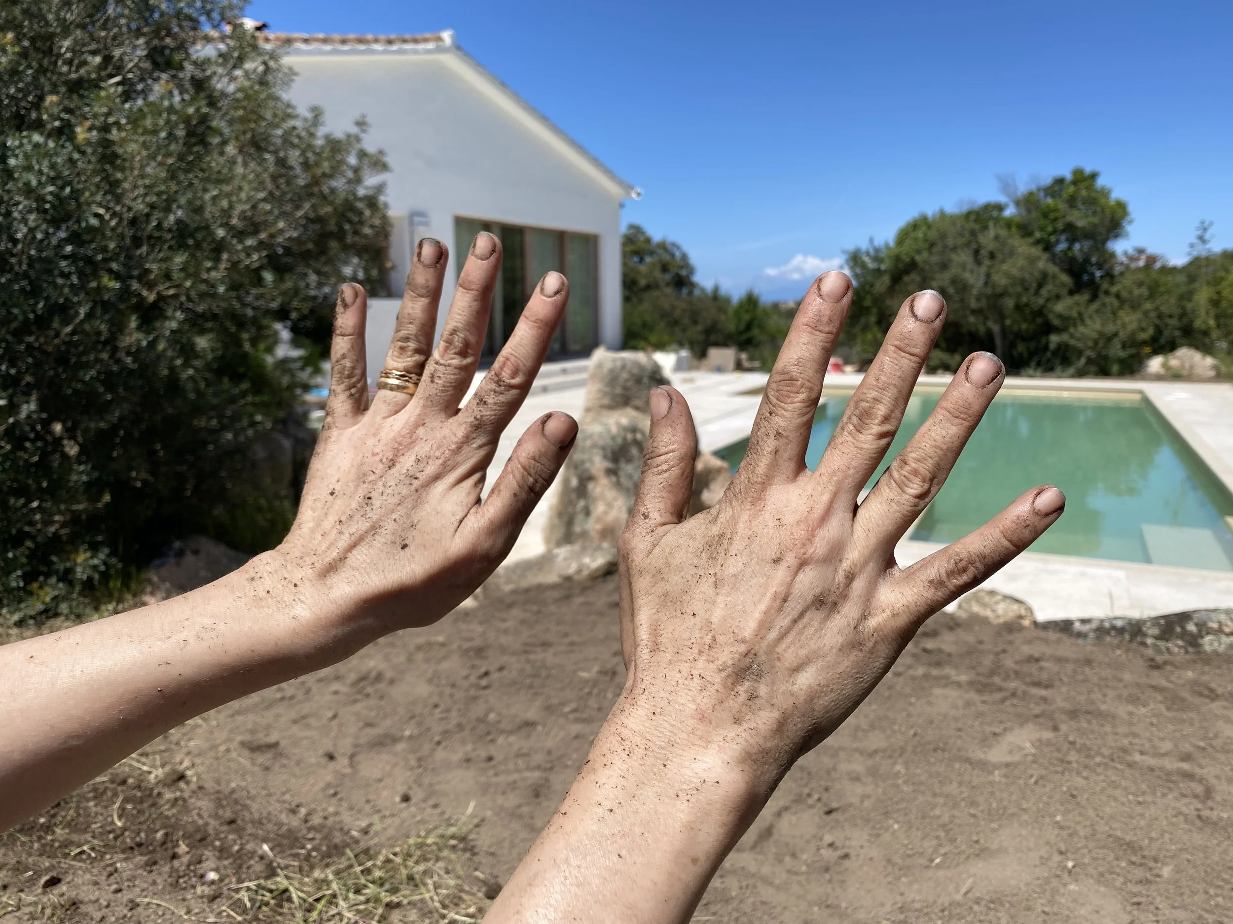 Two hands with dirt and mud on them held up in front of a backyard with a swimming pool, trees, and a white house under a clear blue sky.