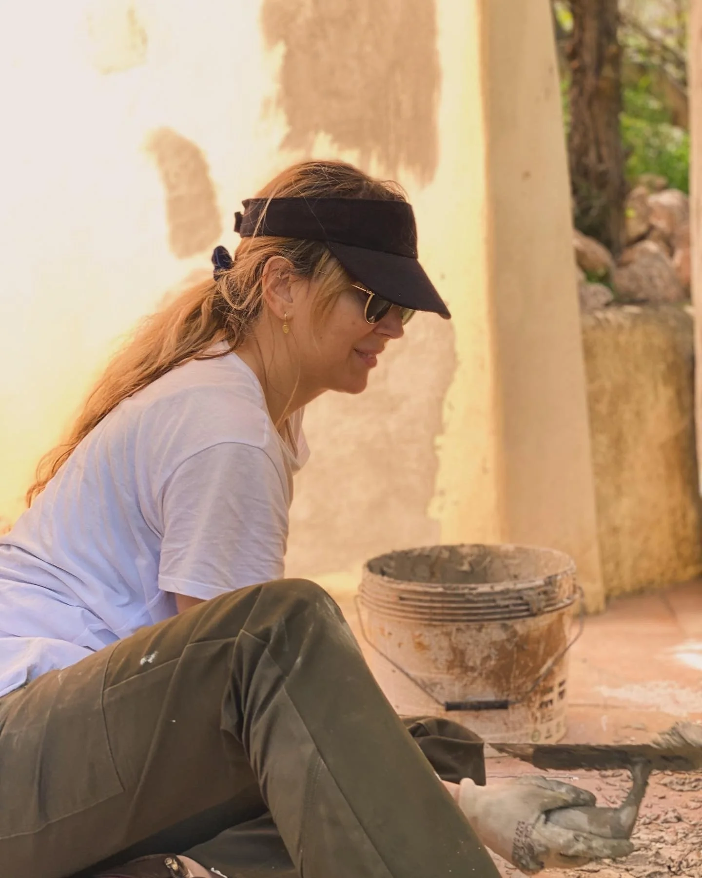 A woman with blonde hair wearing a black visor, sunglasses, a white t-shirt, and brown pants is sitting on the ground, working on a construction or renovation project. She is smiling and using a trowel on the ground near a bucket of mortar or concret