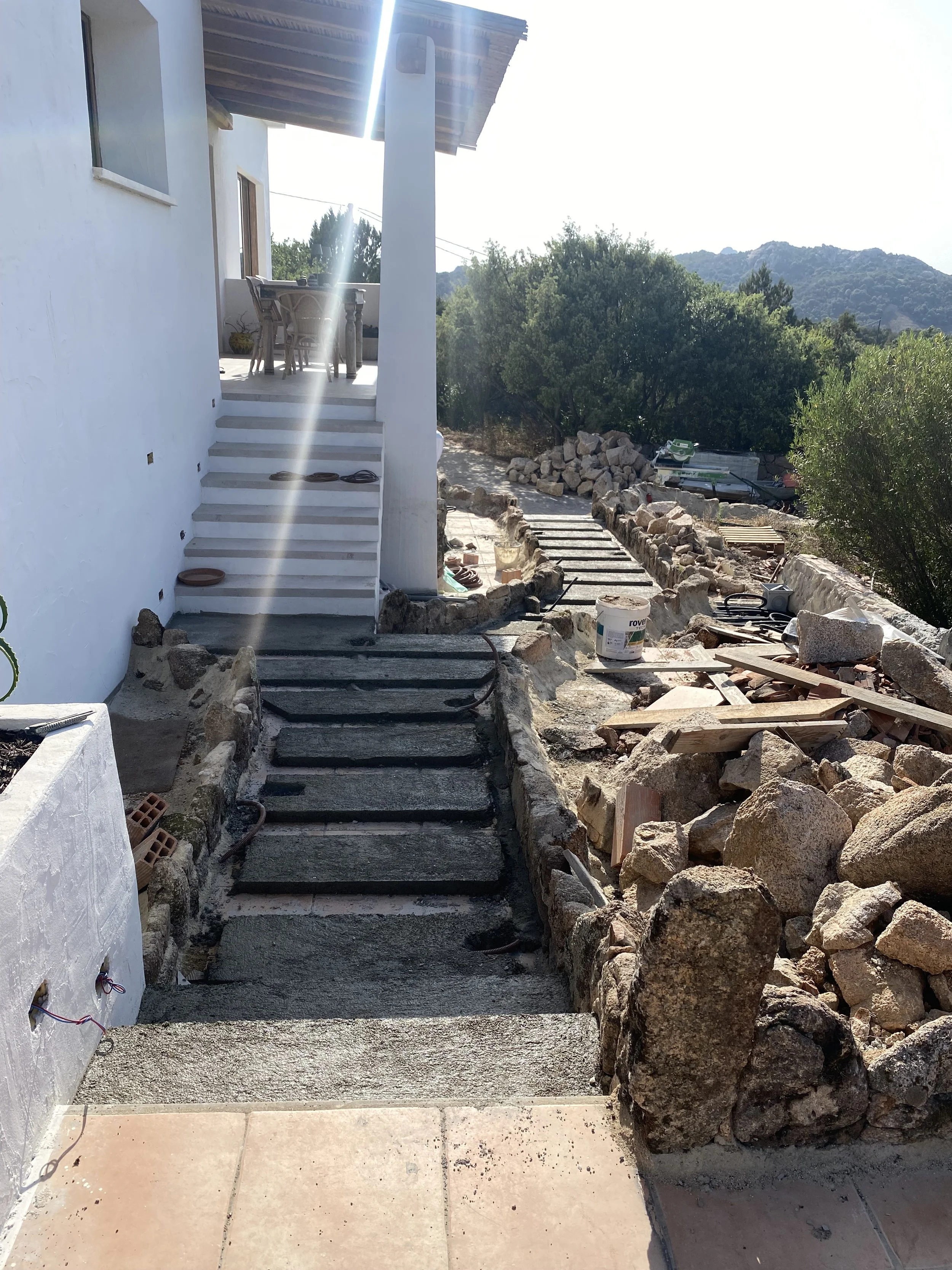 Construction site with stairs leading to an outdoor patio of a white house, surrounded by rocks and construction materials, in a hilly area with trees and mountains in the background.