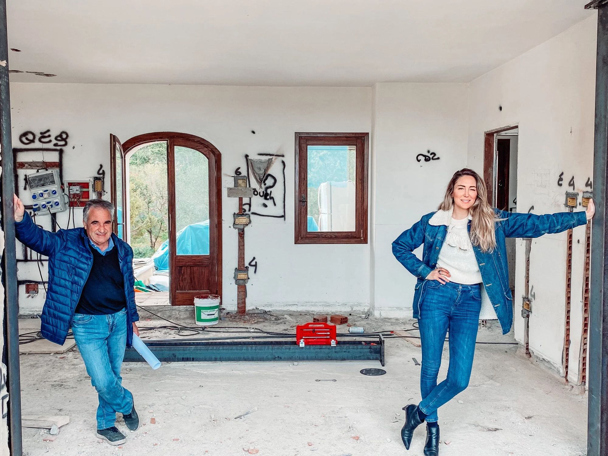 Two people standing inside a house under construction, smiling, with construction tools and electrical wires visible.