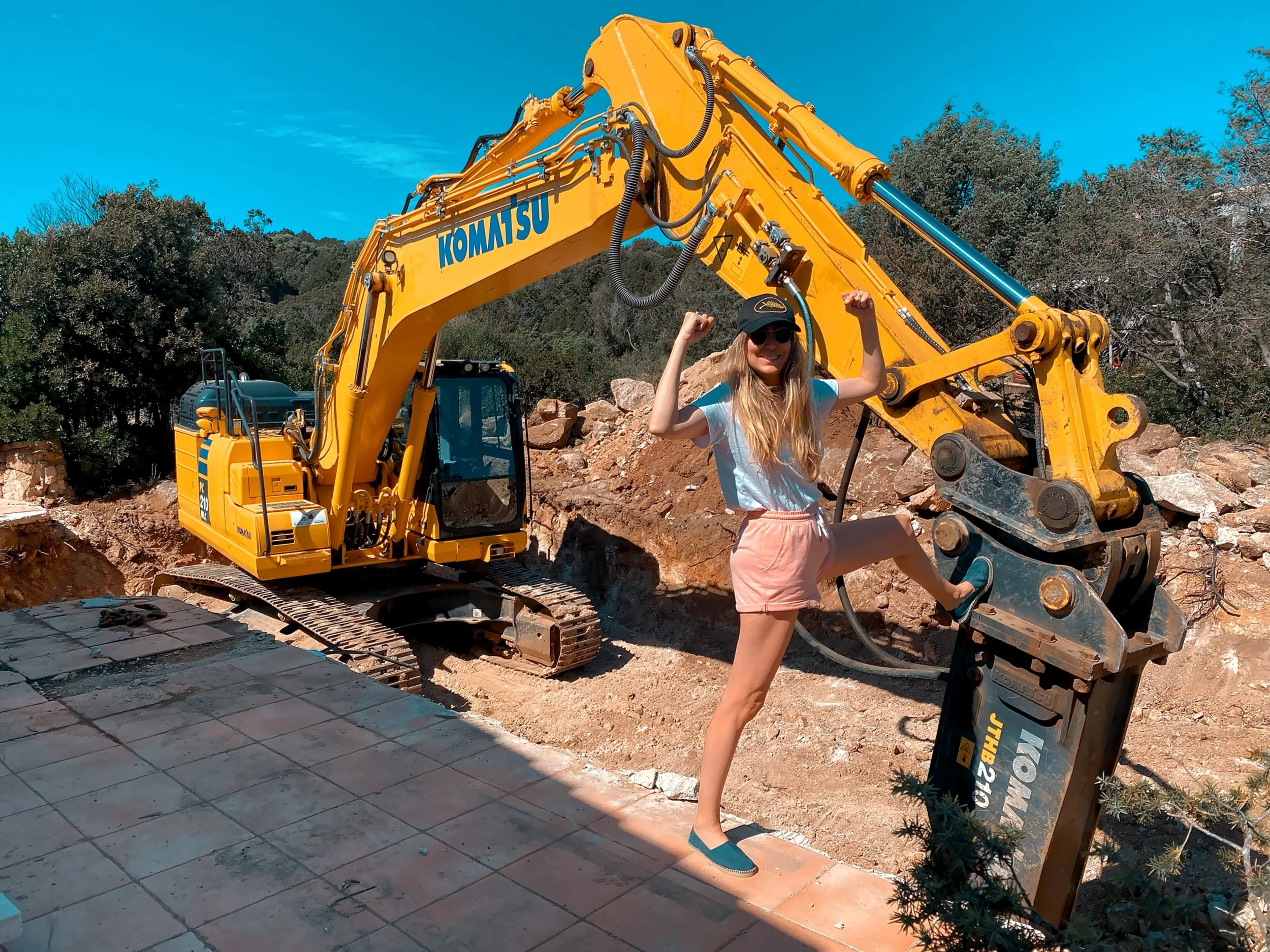 A woman with long blonde hair, wearing sunglasses, a black cap, a white t-shirt, pink shorts, and teal shoes, standing on a tiled surface and flexing her arms. She is in front of a large yellow Komatsu excavator at a construction site with dirt, rock