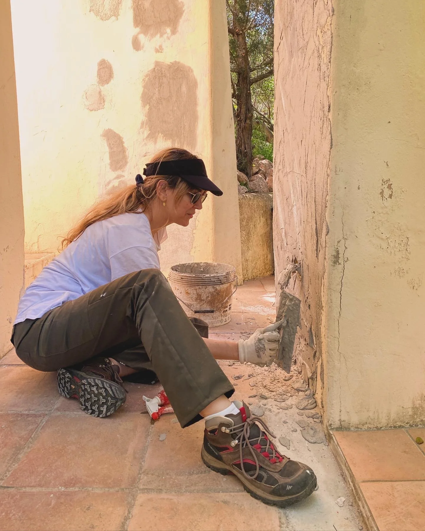 Woman sitting on the floor, fixing a cracked wall with a trowel, wearing a white t-shirt, brown pants, sneakers, a black visor, and sunglasses, with tools and a bucket nearby.