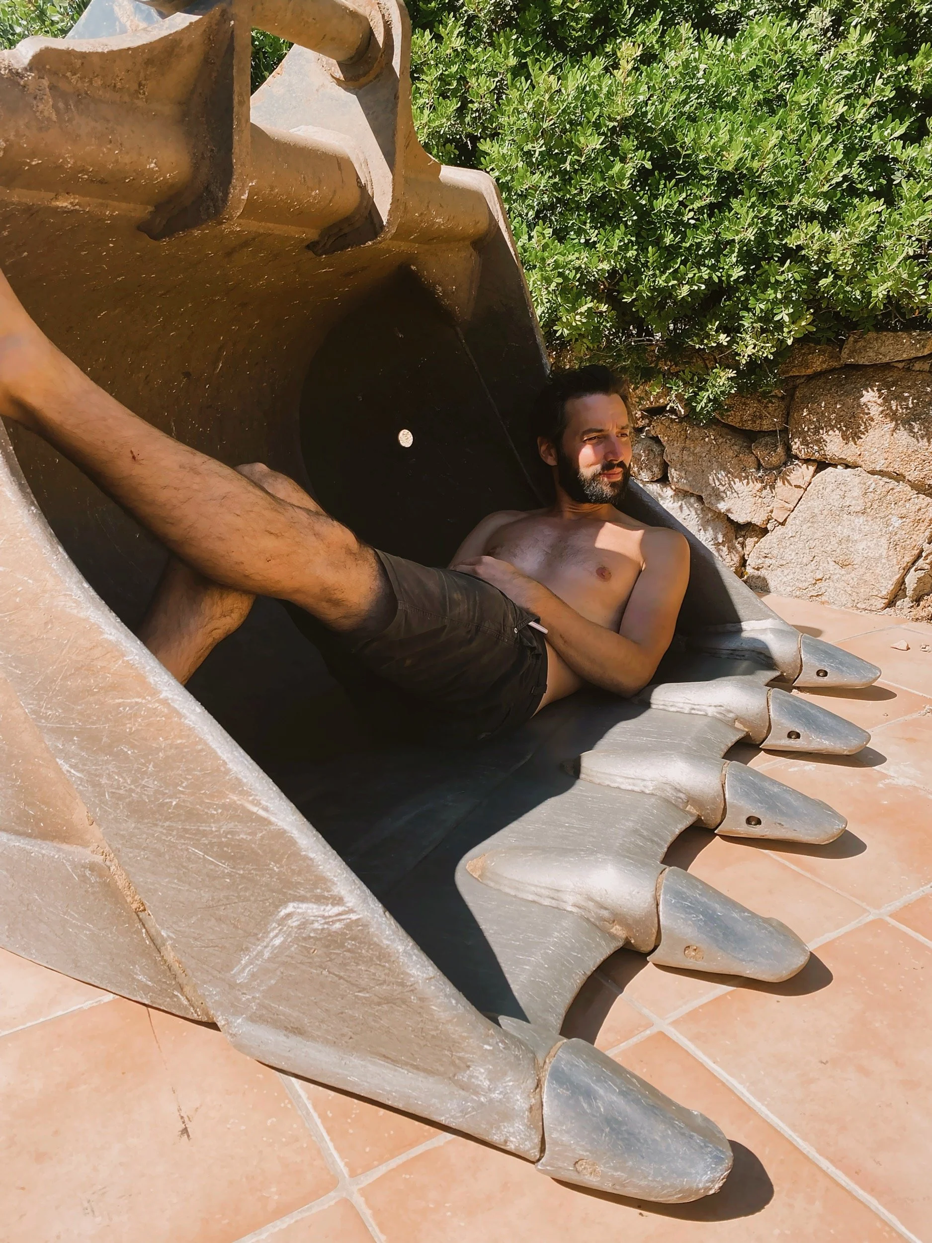 A shirtless man with dark hair and a beard lies on a large, decorative excavation digger bucket, resting on a tiled surface with green bushes and rocks in the background.