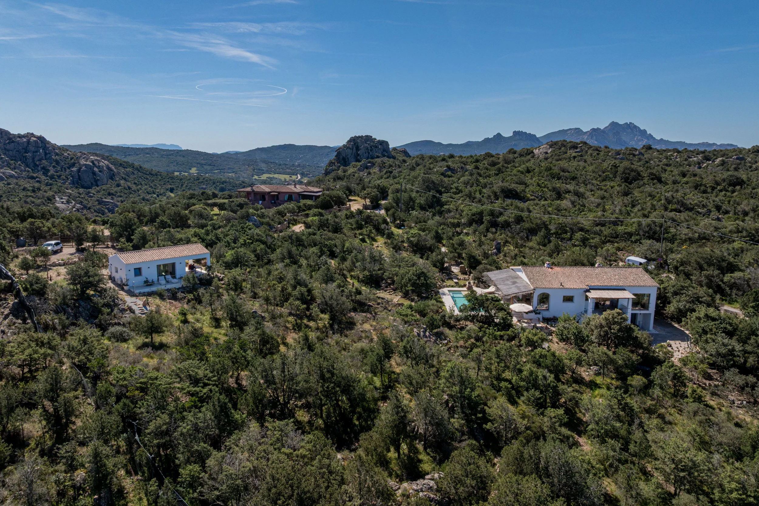 House in Sardinia, Costa Smeralda, with swimming pool surrounded by trees and outdoor furniture, with a rock and stone pathway in the foreground.