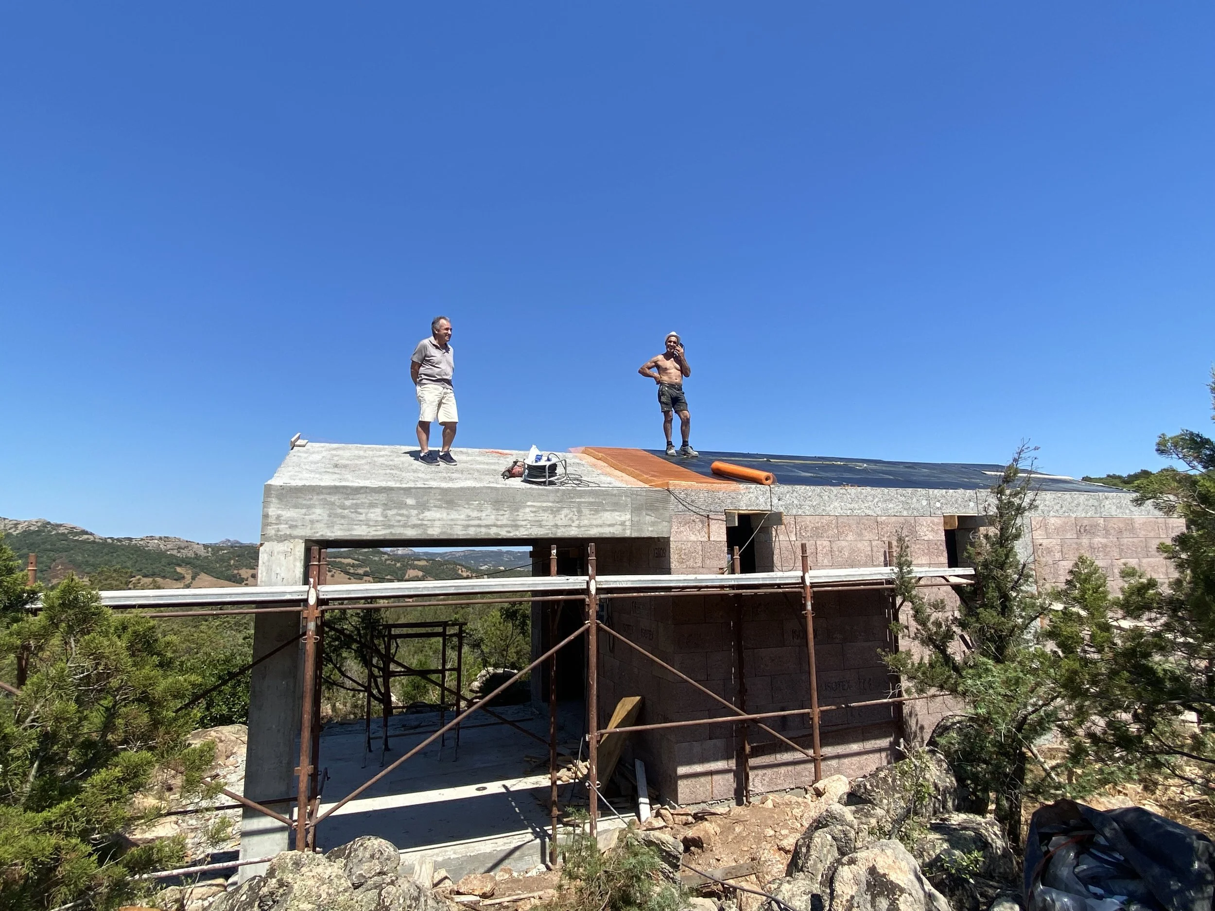 Two construction workers on a partially built concrete structure with a sloped roof, outdoors on a sunny day, surrounded by trees and rocks.