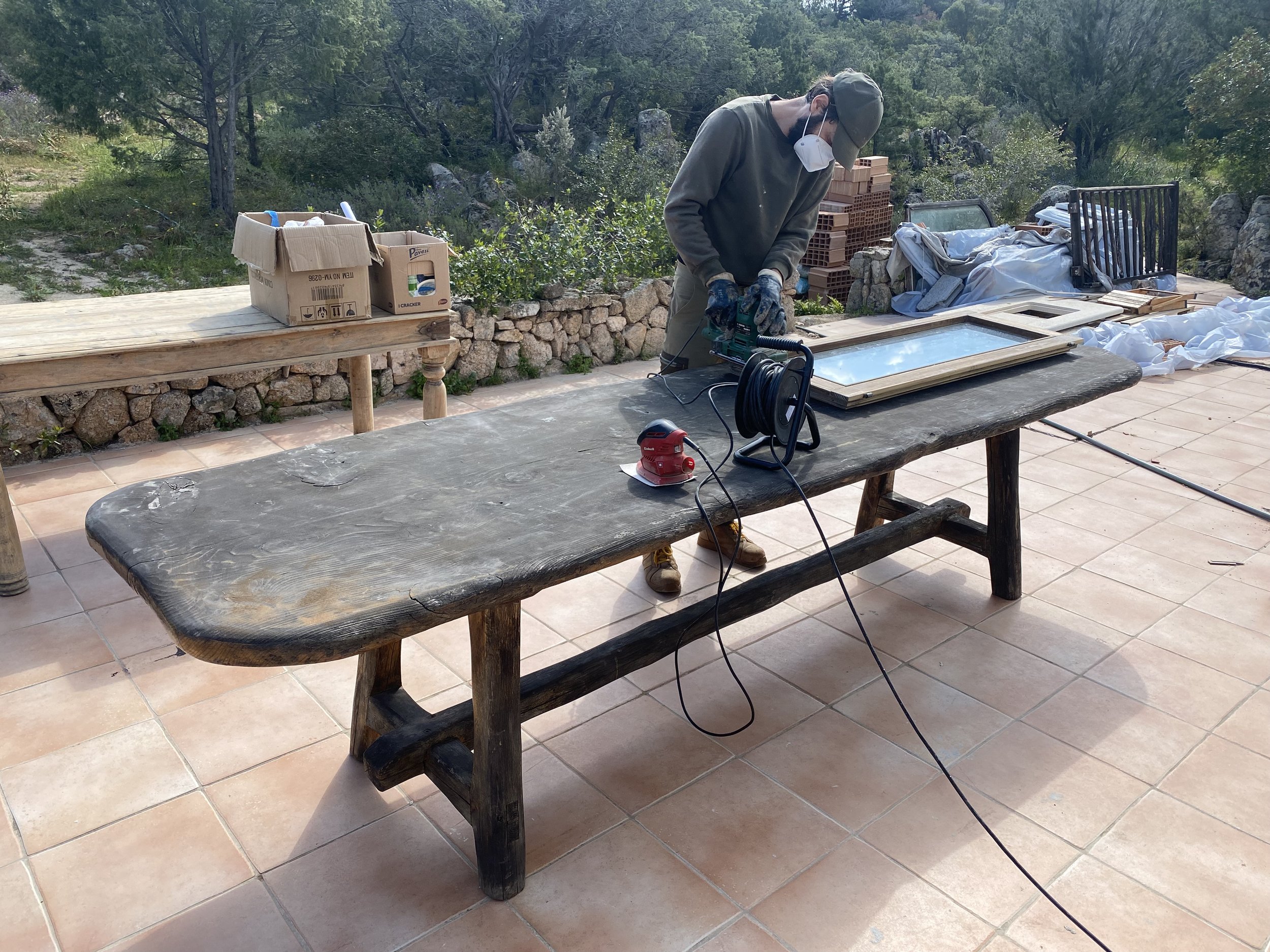 A person wearing a mask, gloves, and a cap is using a power tool on a wooden picture frame on an outdoor table. The scene appears to be a workshop or construction area with other work materials and supplies nearby, set against a backdrop of greenery.