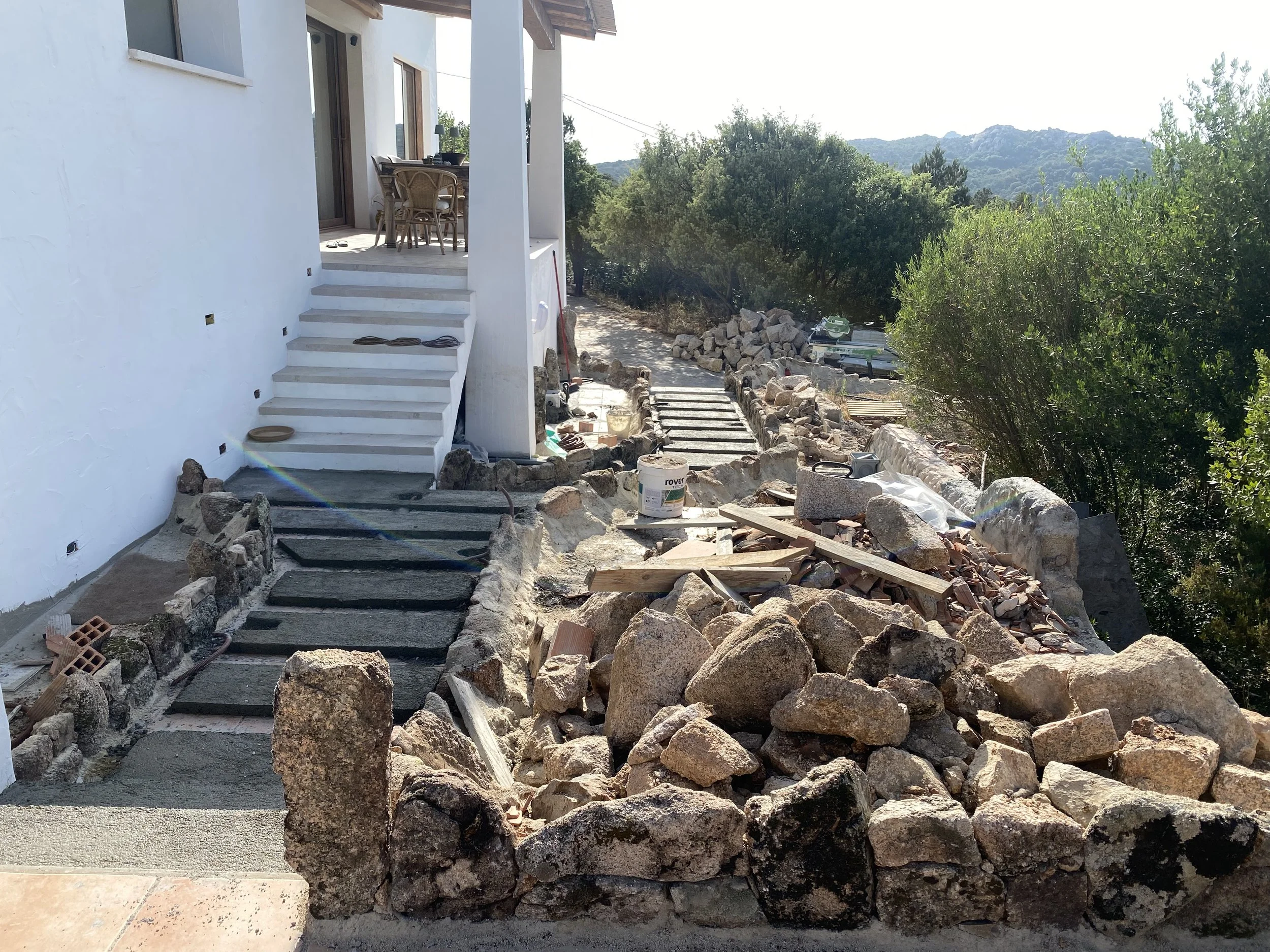Construction of an outdoor stone pathway leading to a white house with stairs, with construction materials and rocks scattered around, and a view of trees and hills in the background.